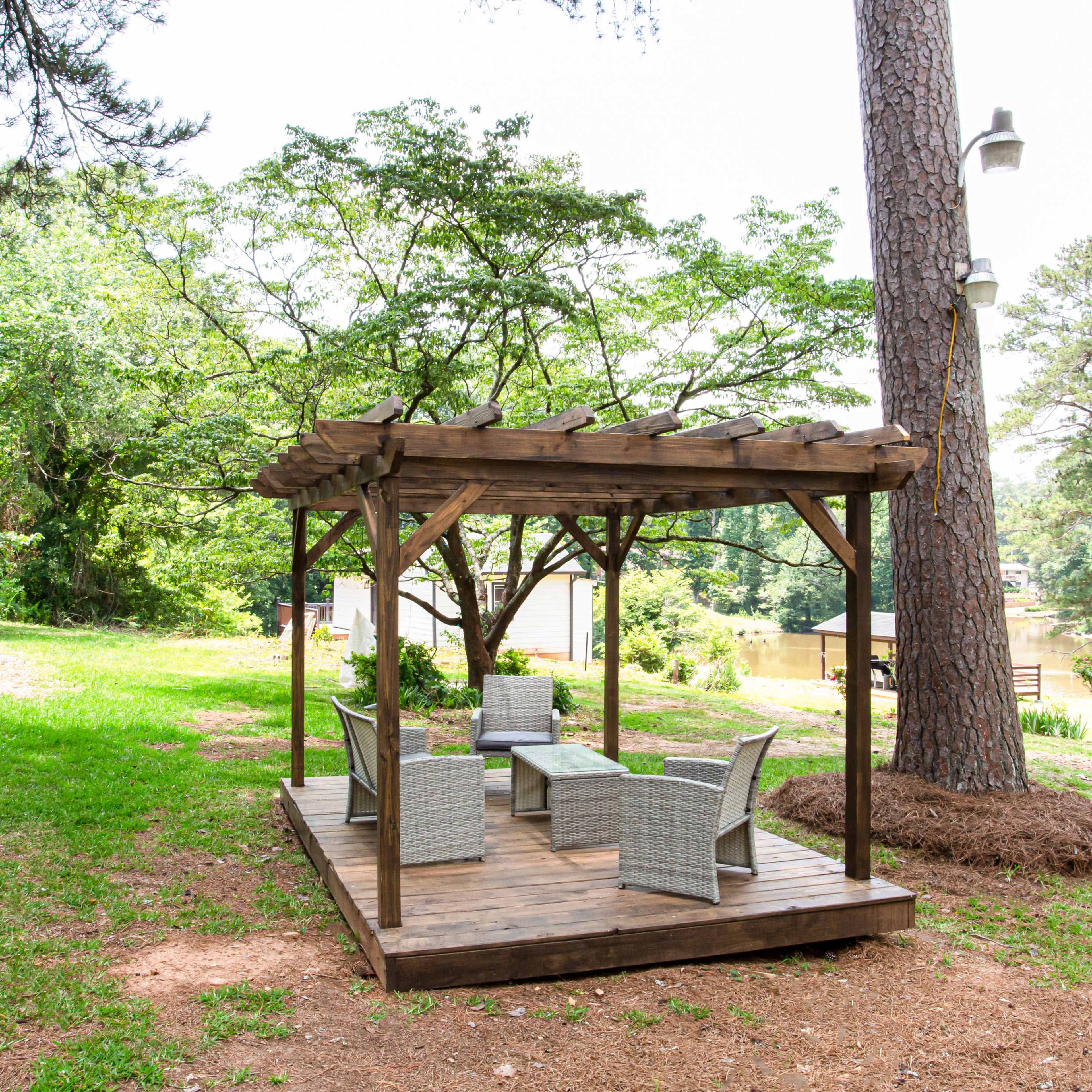 A wooden gazebo with four chairs and a table is set on a grassy area near a river, surrounded by trees.