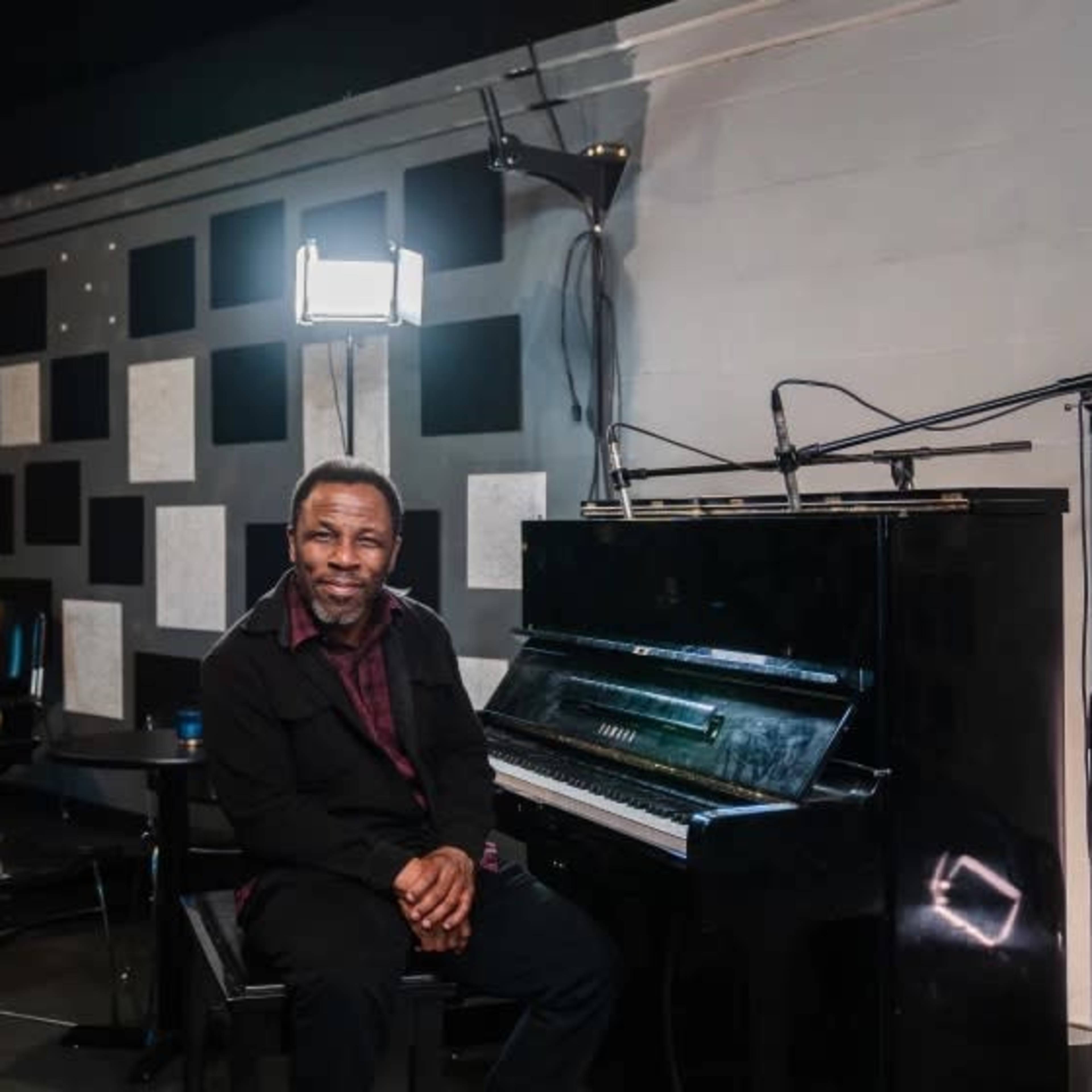 A musician sits beside a black piano in a dimly lit studio with geometric wall panels.
