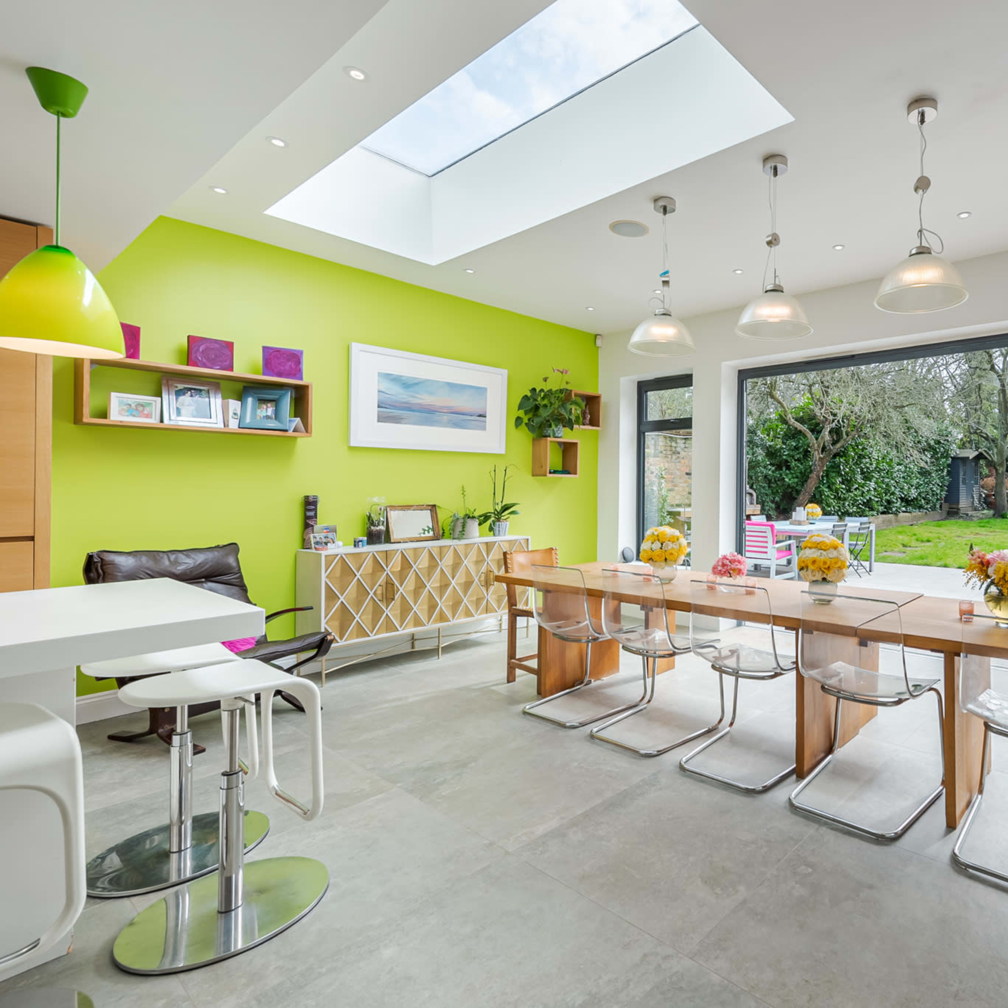 A modern kitchen features a long wooden dining table with clear chairs, large glass doors opening to a garden, and a bright green accent wall.