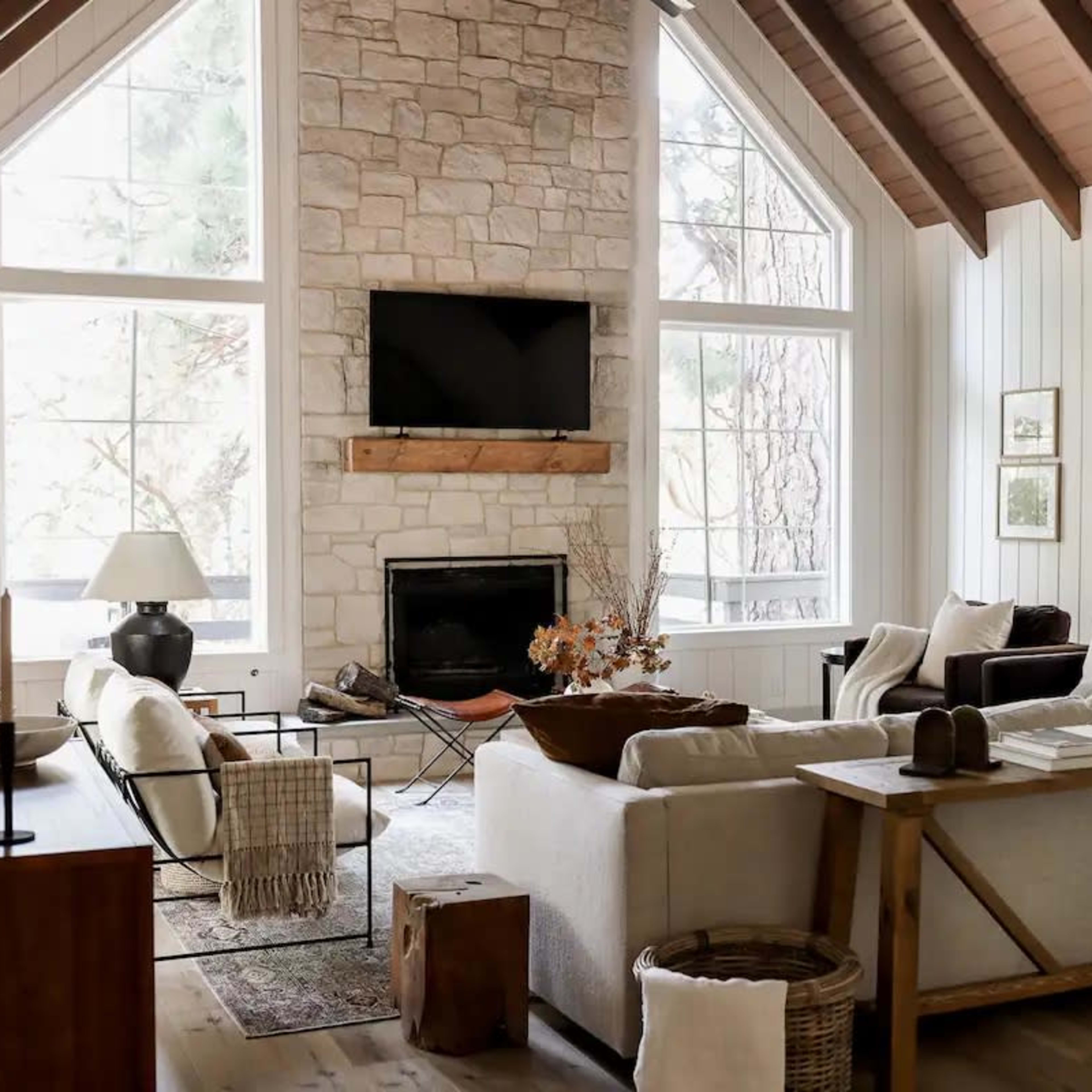 A bright living room featuring a stone fireplace, large windows, and a mix of modern and rustic furniture arrangements.