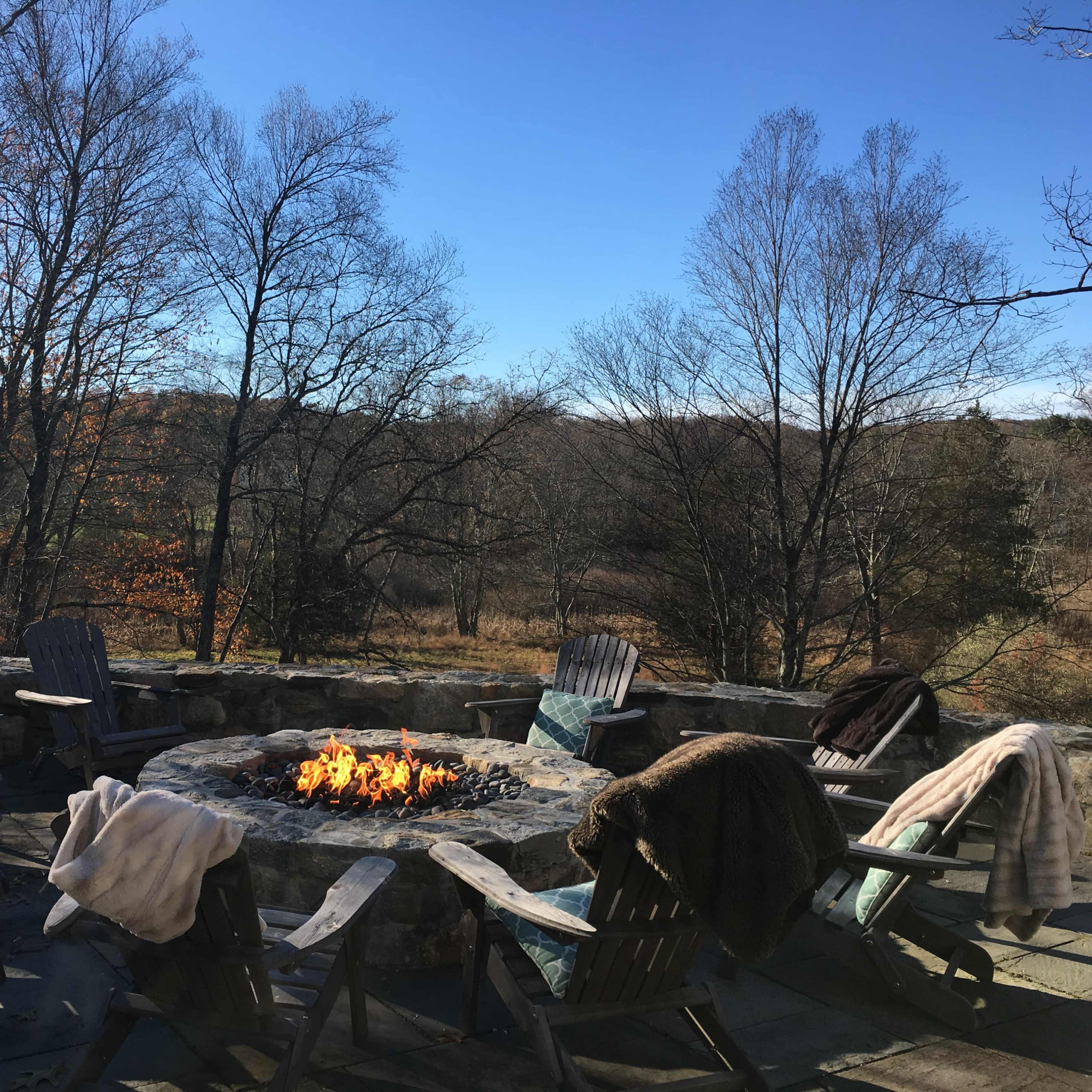 A stone fire pit surrounded by wooden chairs and blankets sits in a clearing with bare trees and a blue sky in the background.