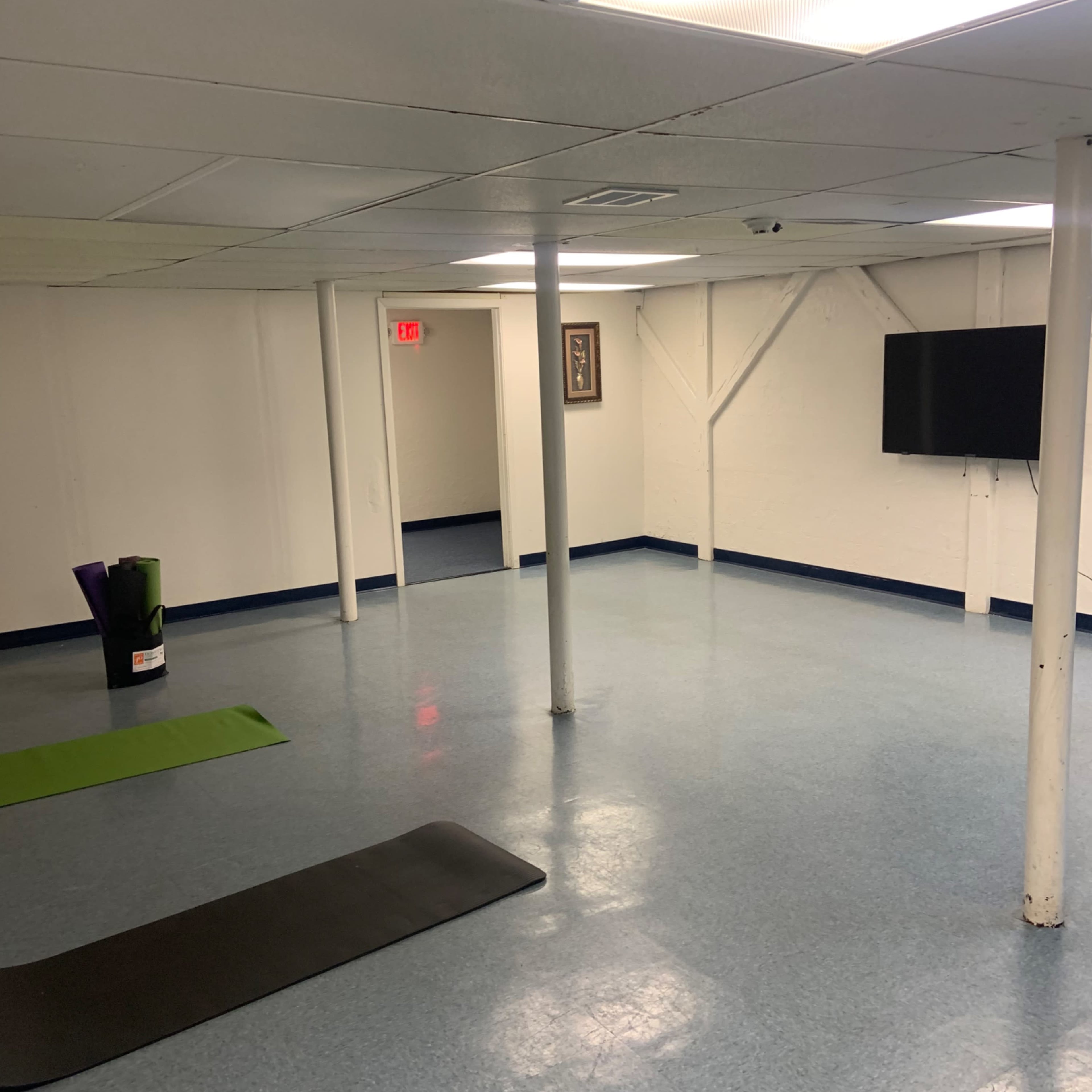 A spacious, empty basement room with blue flooring, a yoga mat, a green exercise mat, a television mounted on the wall, and structural support poles.