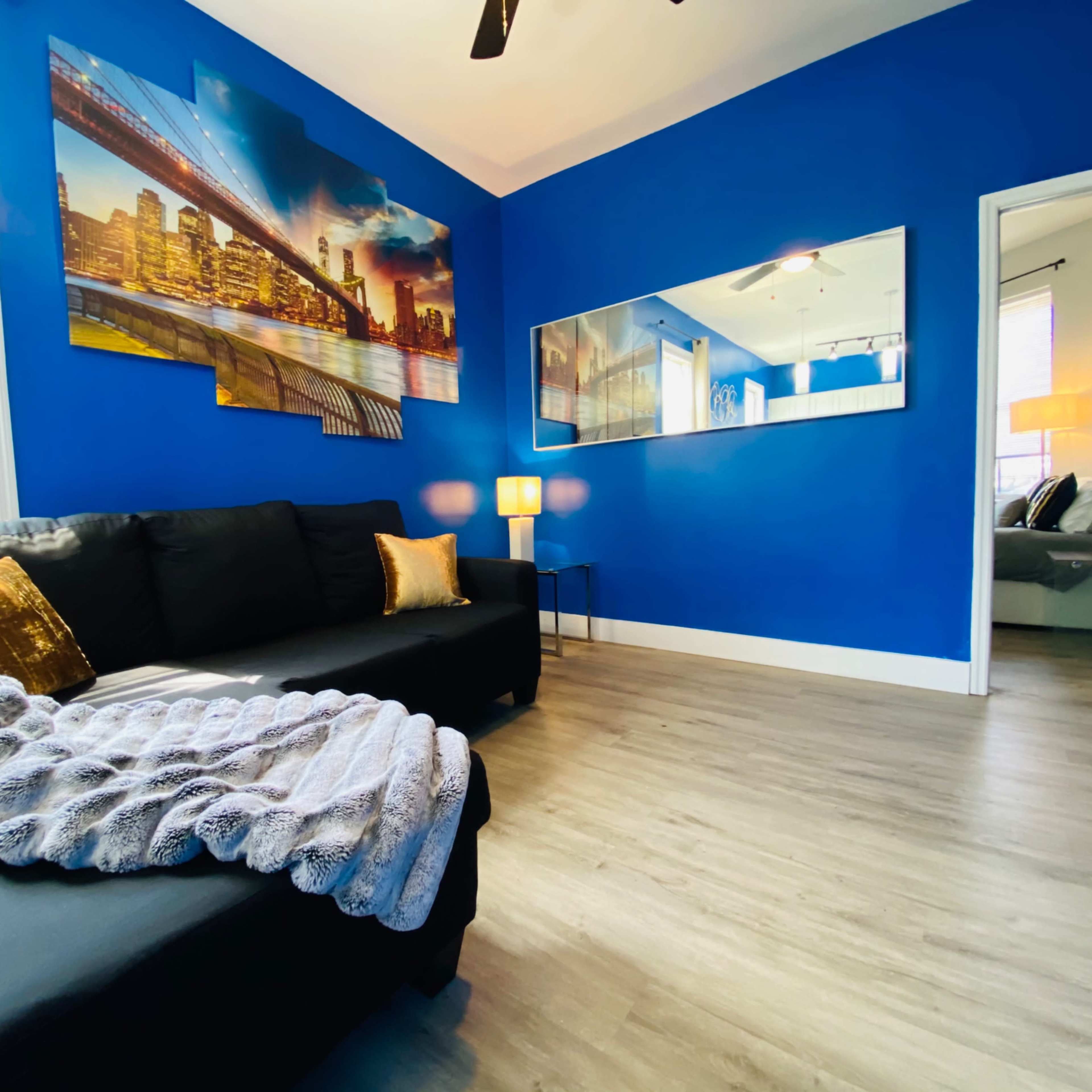 A living room with a blue wall, a black sofa adorned with a gray throw blanket, and a large cityscape painting above it, complemented by two lamps and a doorway leading to another room.