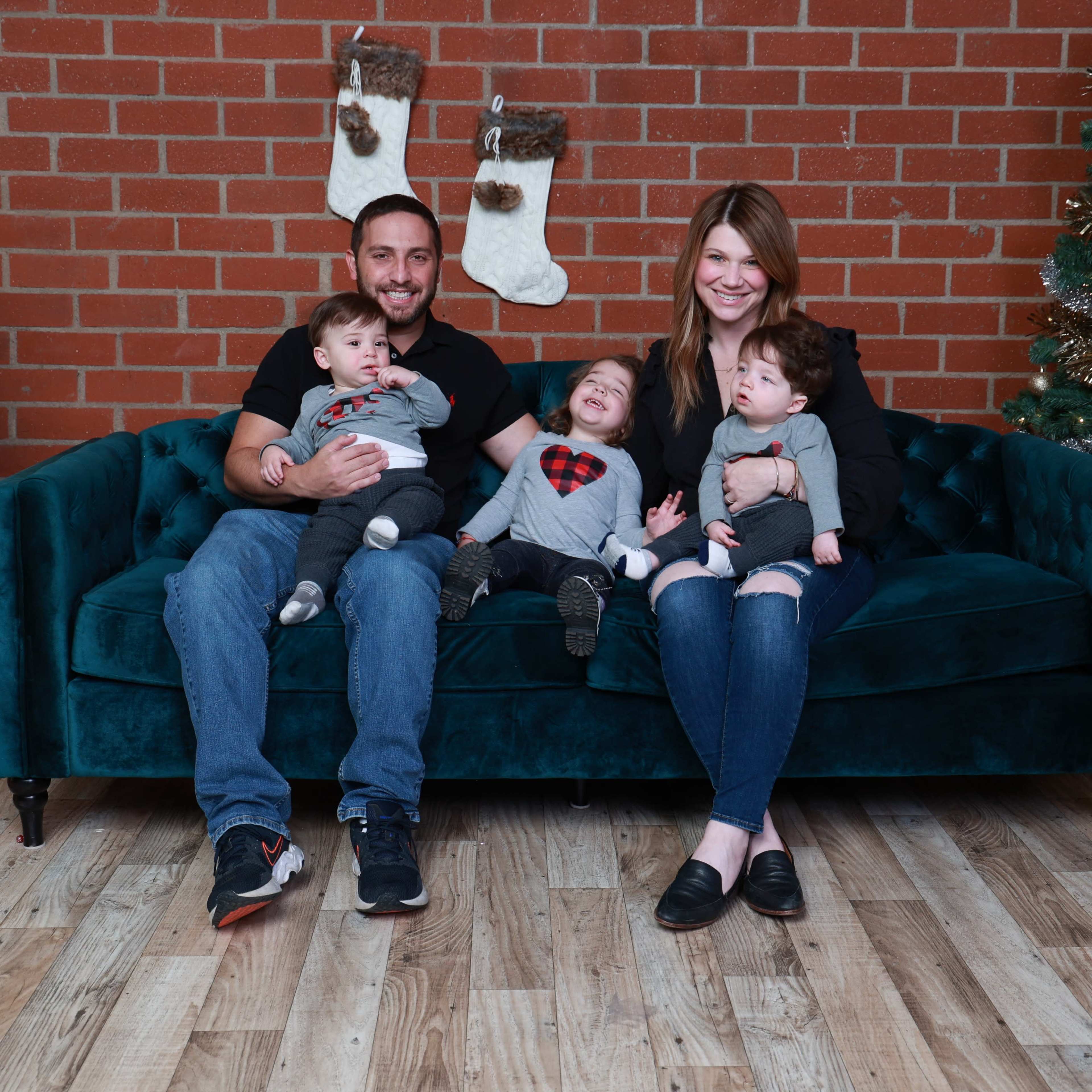 A family of four sits on a teal couch in front of a decorated Christmas tree and holiday presents.