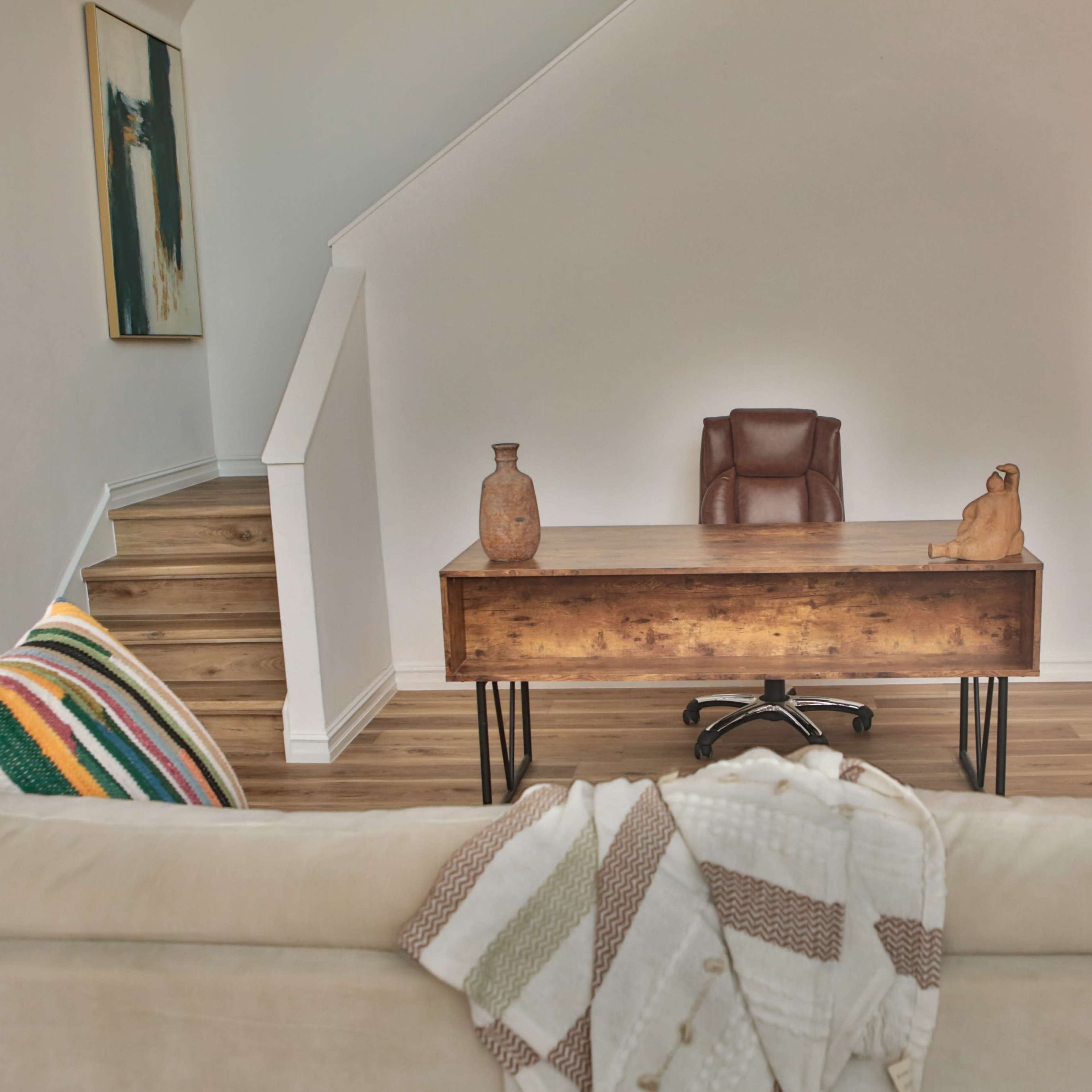 A wooden desk with a leather chair is positioned in an airy room, adjacent to a staircase and decorated with two pottery pieces on the desk.