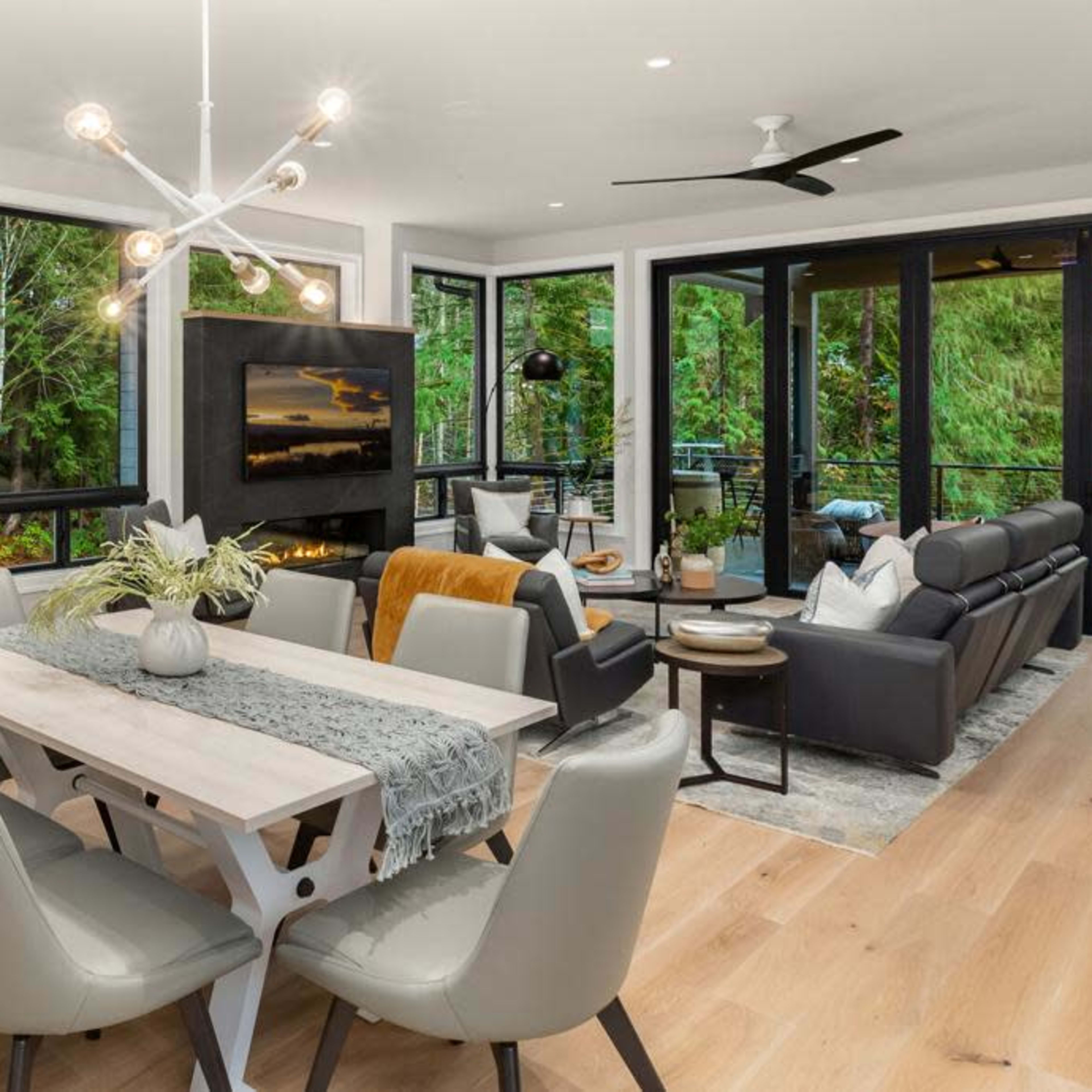 A modern living and dining area features a dining table with chairs, a seating arrangement around a fireplace, and large windows overlooking greenery.