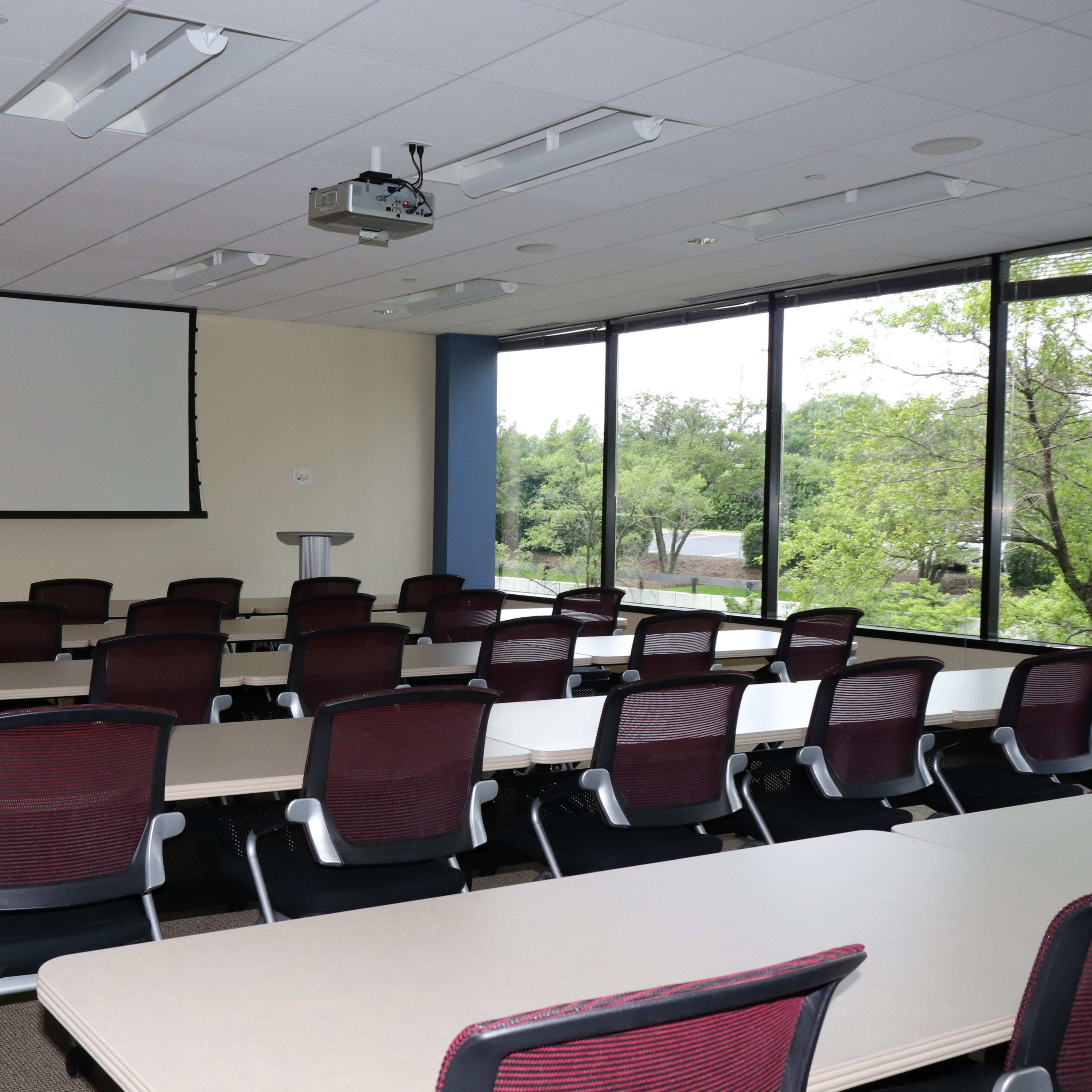 A conference room with multiple tables and chairs arranged for a meeting, featuring a projector screen and large windows providing natural light.