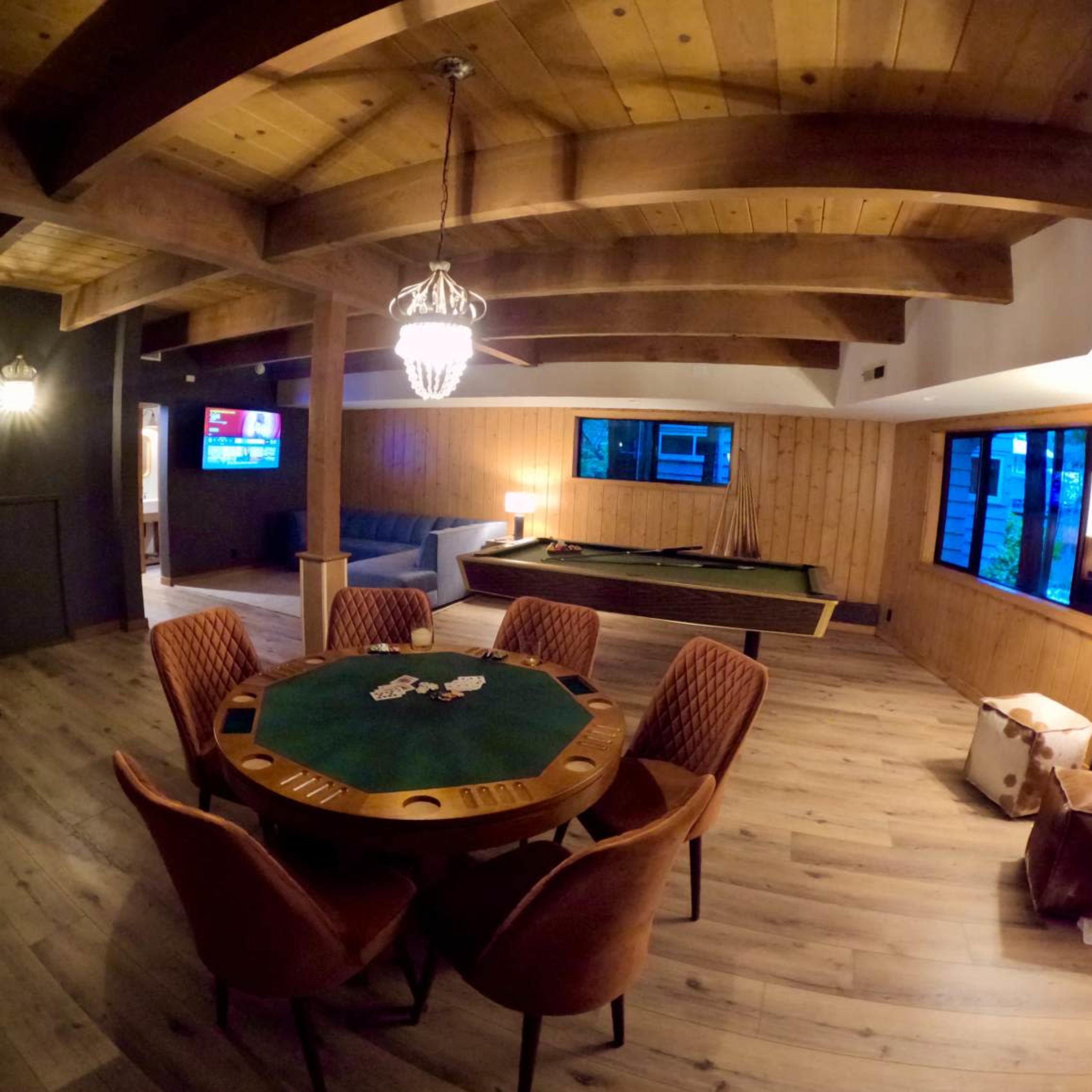 A spacious game room featuring a poker table surrounded by upholstered chairs, a pool table, and large windows allowing natural light to illuminate the wooden interior.