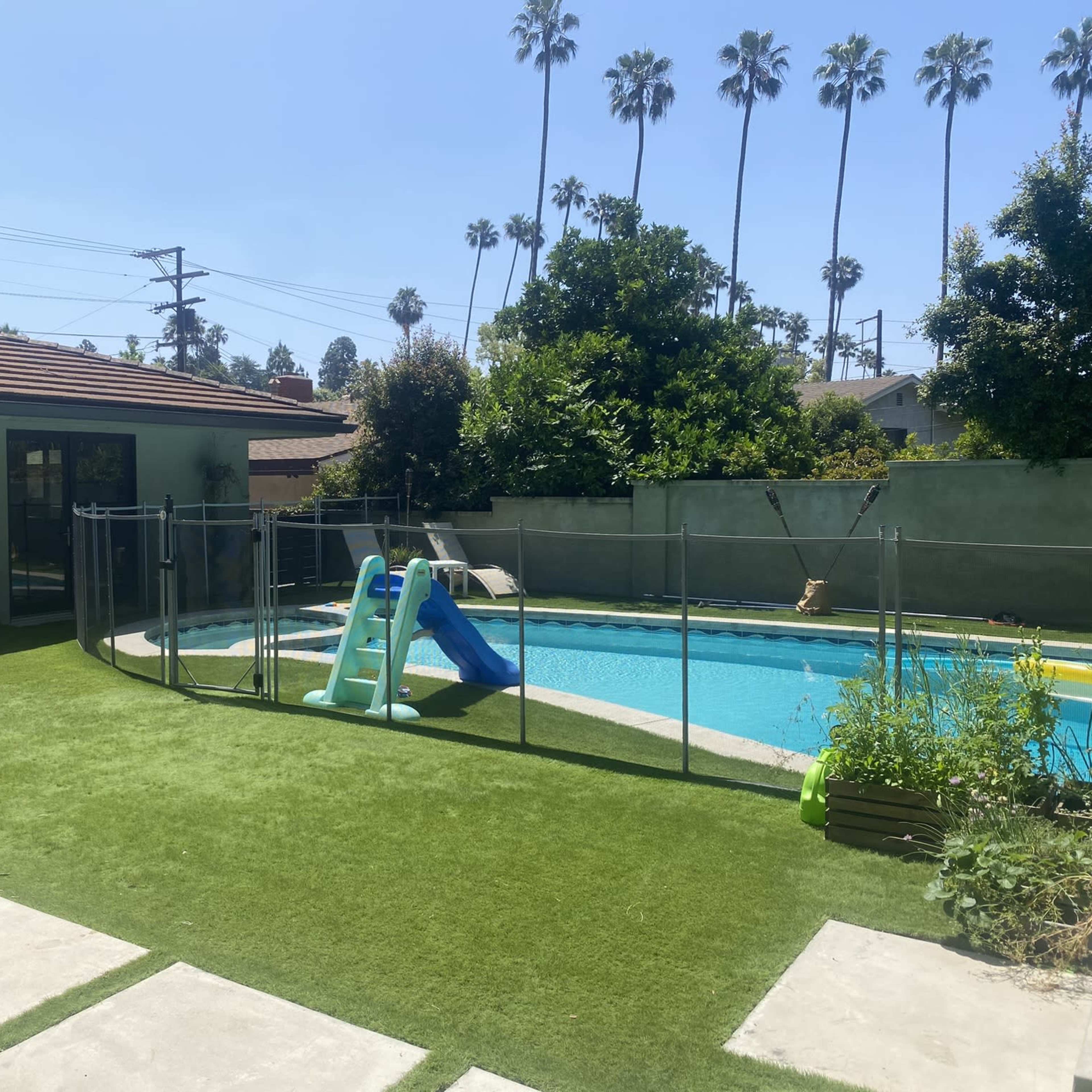 A backyard with a swimming pool, a slide, a lush lawn, and palm trees in the background.