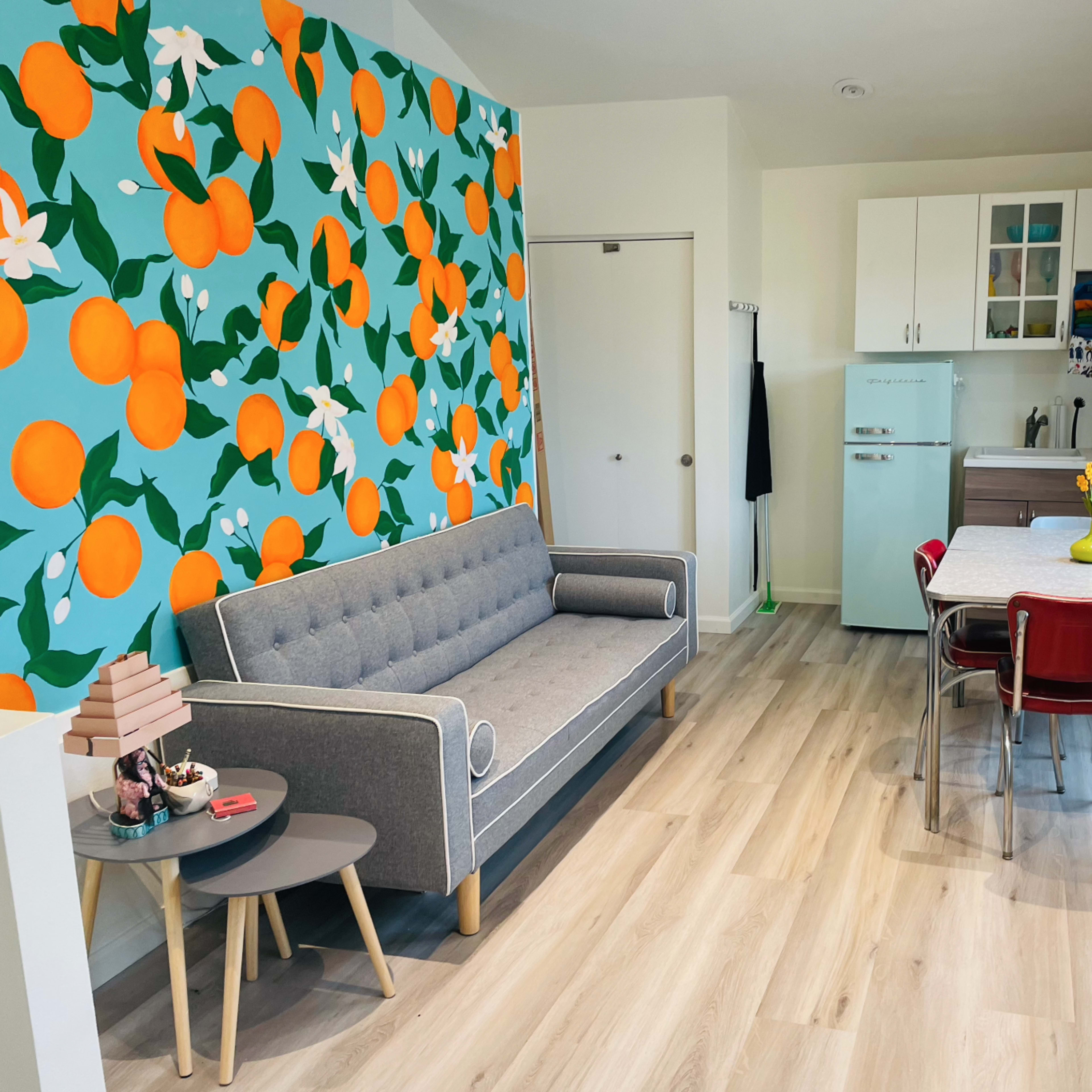 A modern living space with a couch, a dining table with chairs, and a kitchen area, all enhanced by a vibrant orange and floral accent wall.
