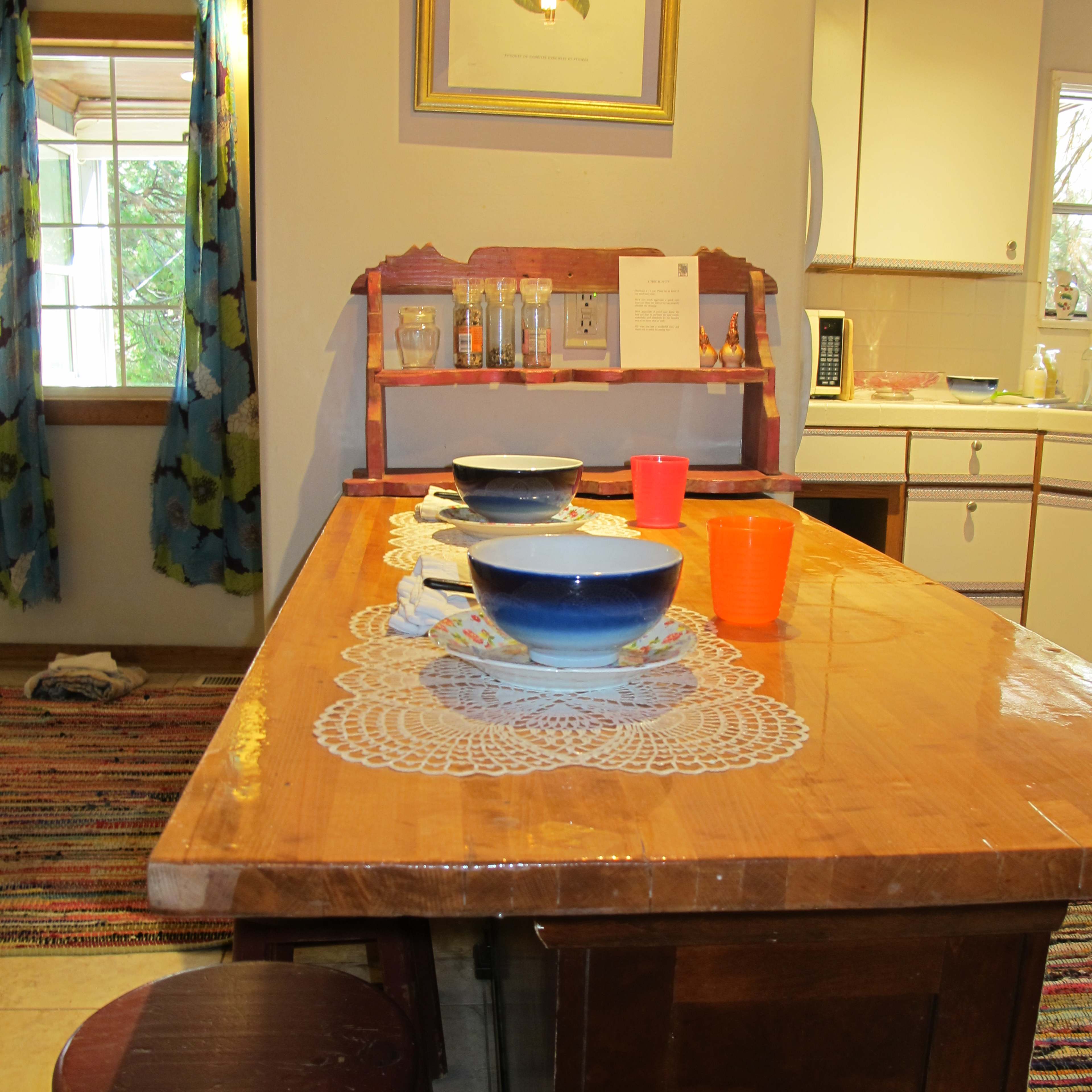 A wooden table is set with bowls and colorful cups in a brightly lit kitchen featuring a large window and a rug.