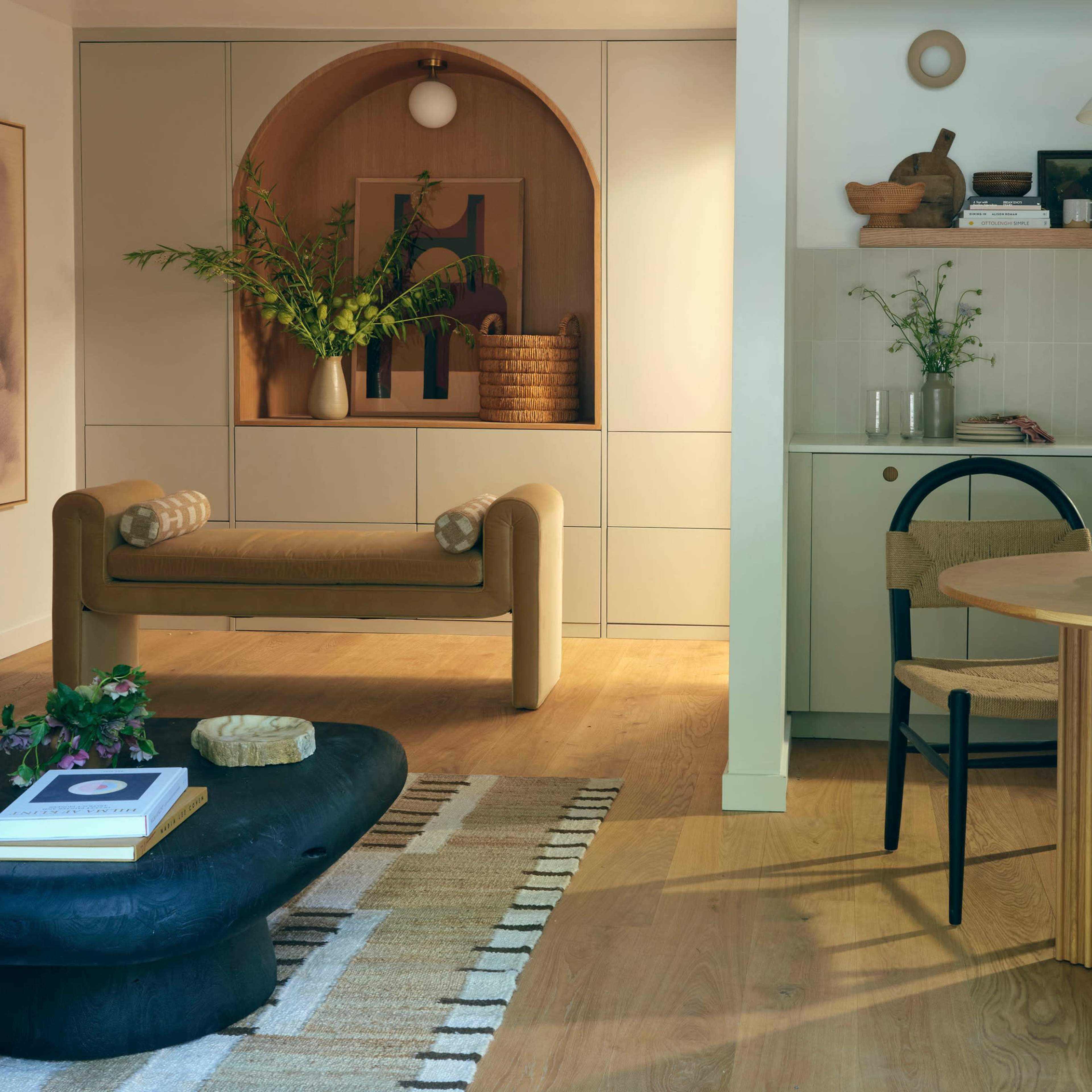 A modern living space featuring a beige sofa, a rounded wooden bench, a circular dining table, and a kitchen area with light wood accents and decorative plants.