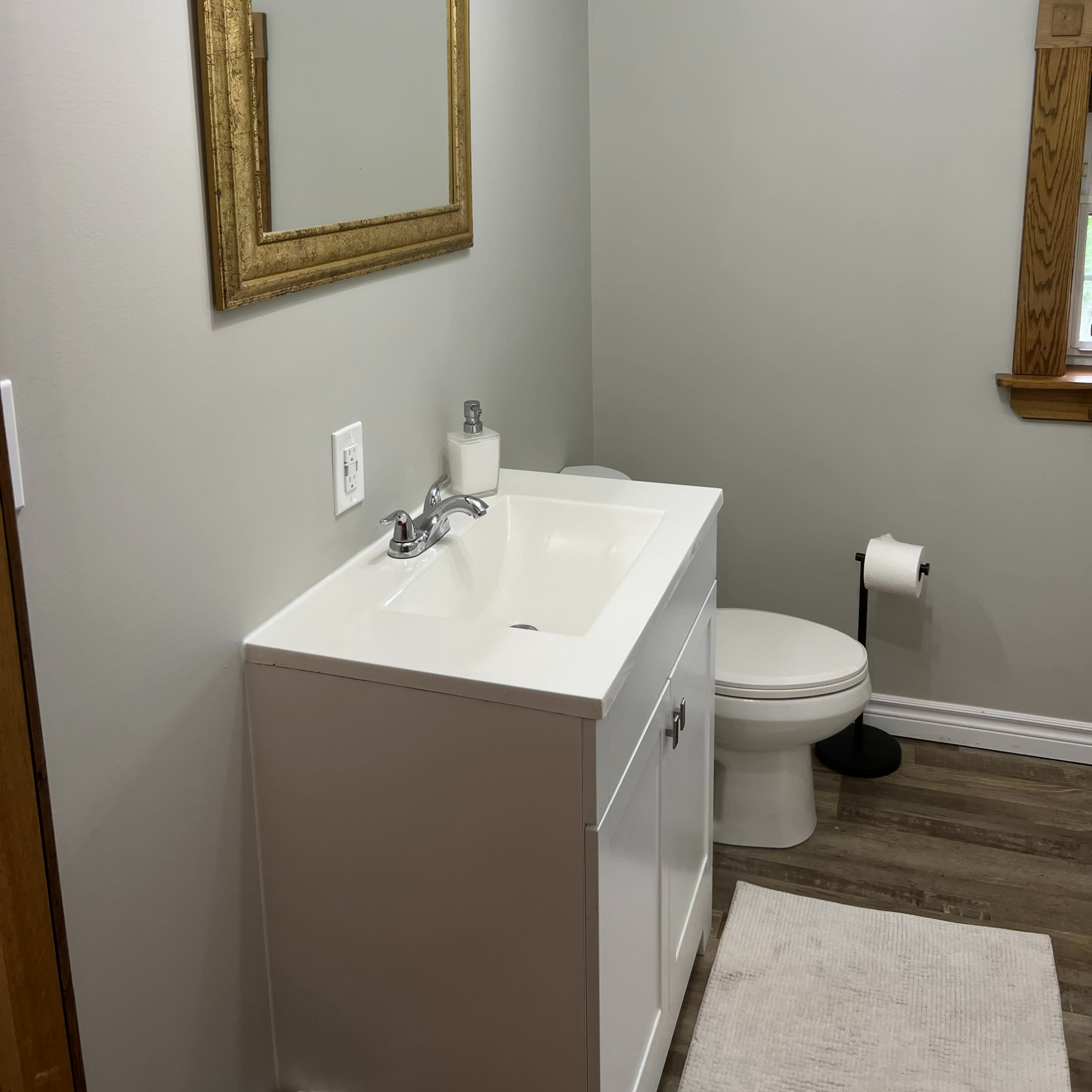 A bathroom featuring a white vanity with a sink, a gold-framed mirror above it, a white toilet, and a window with wooden trim.
