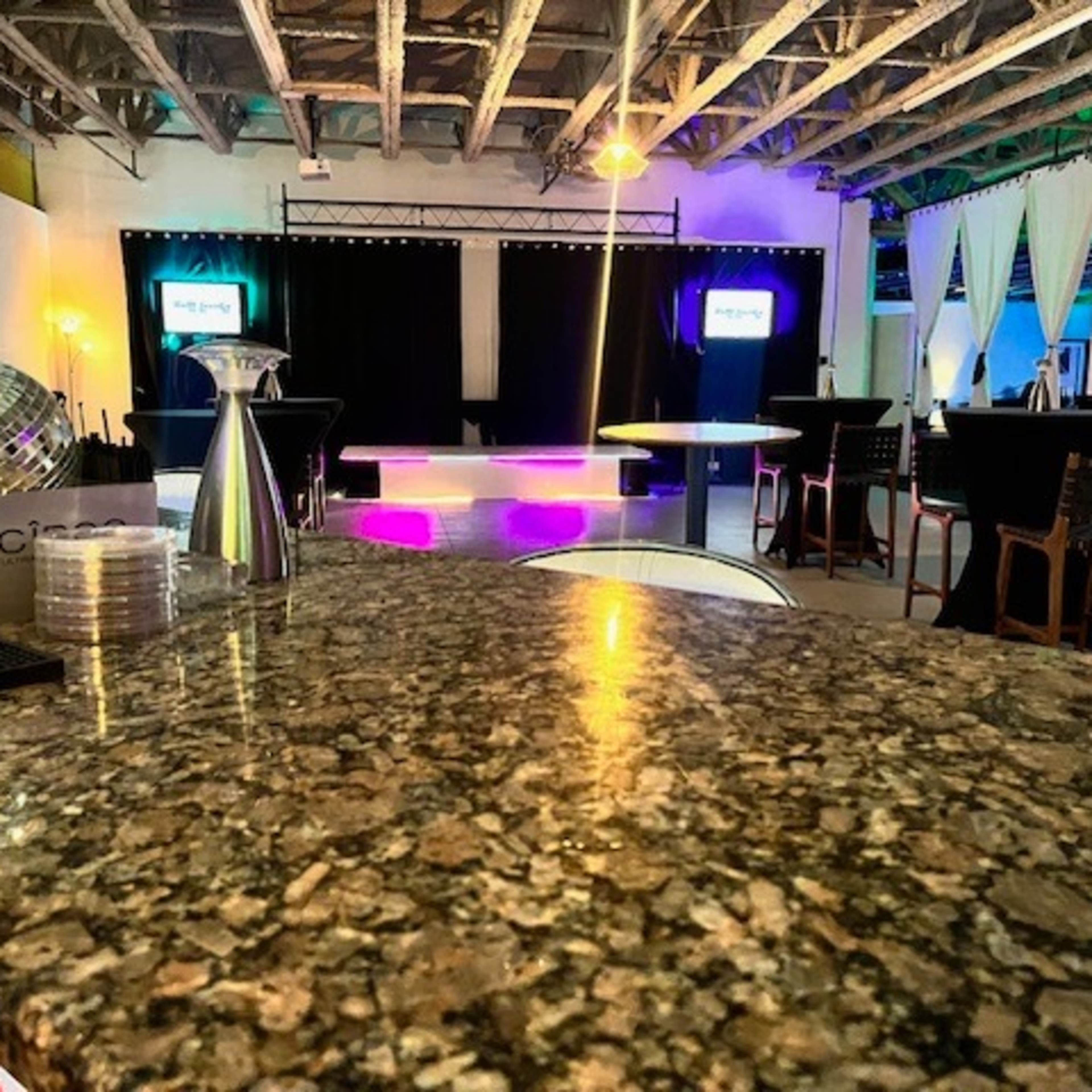 A spacious event venue with a polished granite bar in the foreground, a stage with lights in the background, and several tables and chairs arranged throughout.