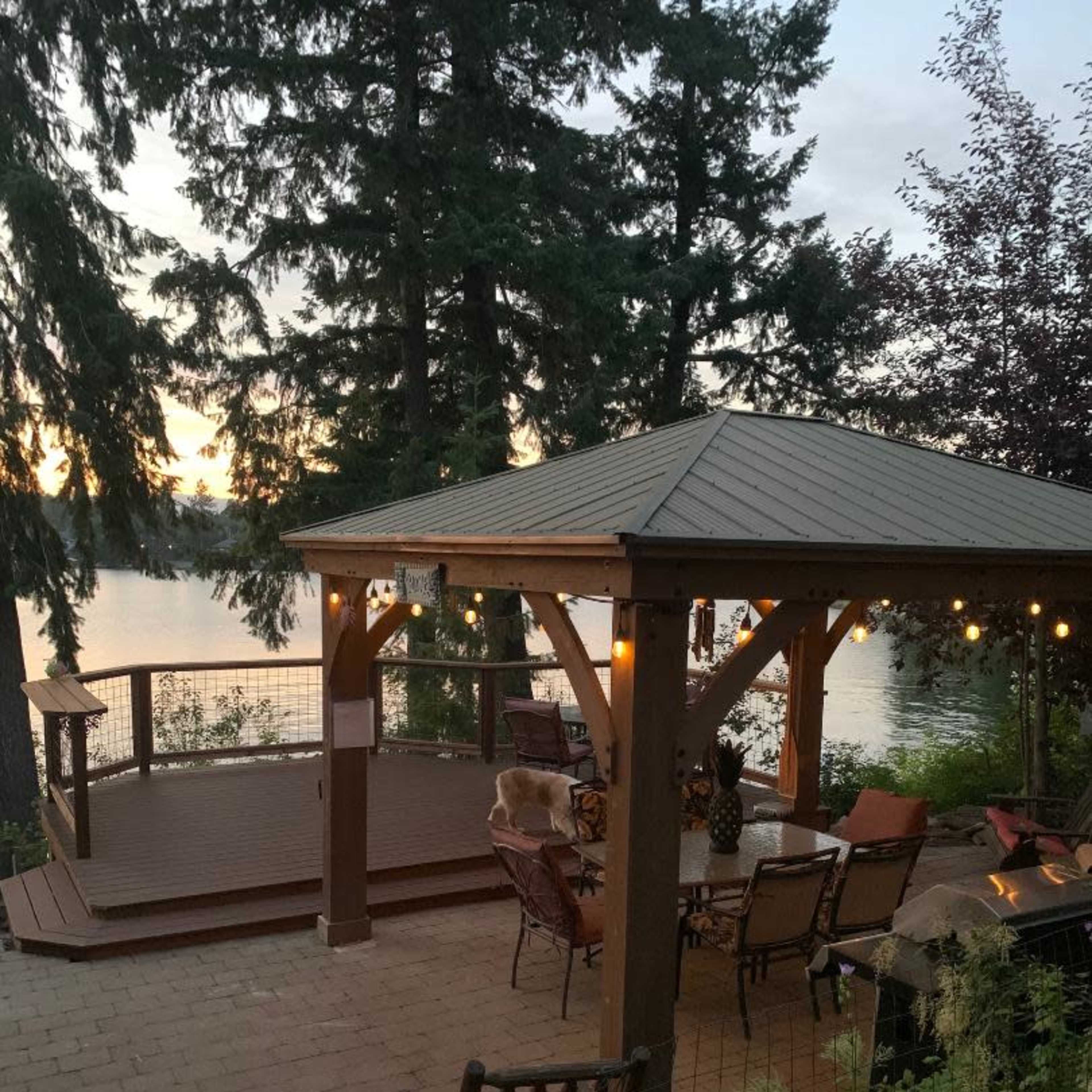A wooden gazebo with string lights overlooks a patio area near a lake, surrounded by tall trees at sunset.