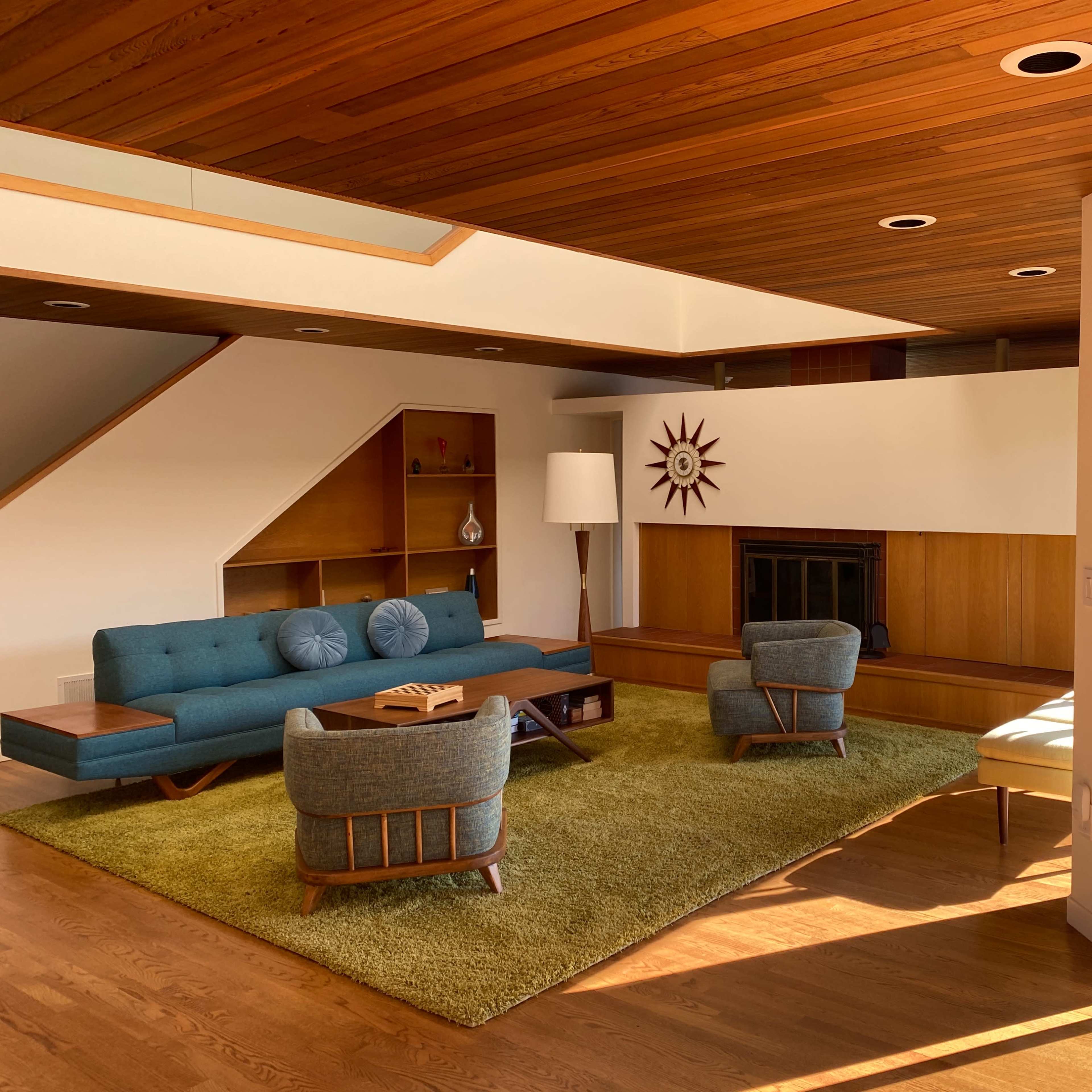 A mid-century modern living room features a teal sofa, two armchairs, a sunburst clock, and large windows opening to a wooden deck.