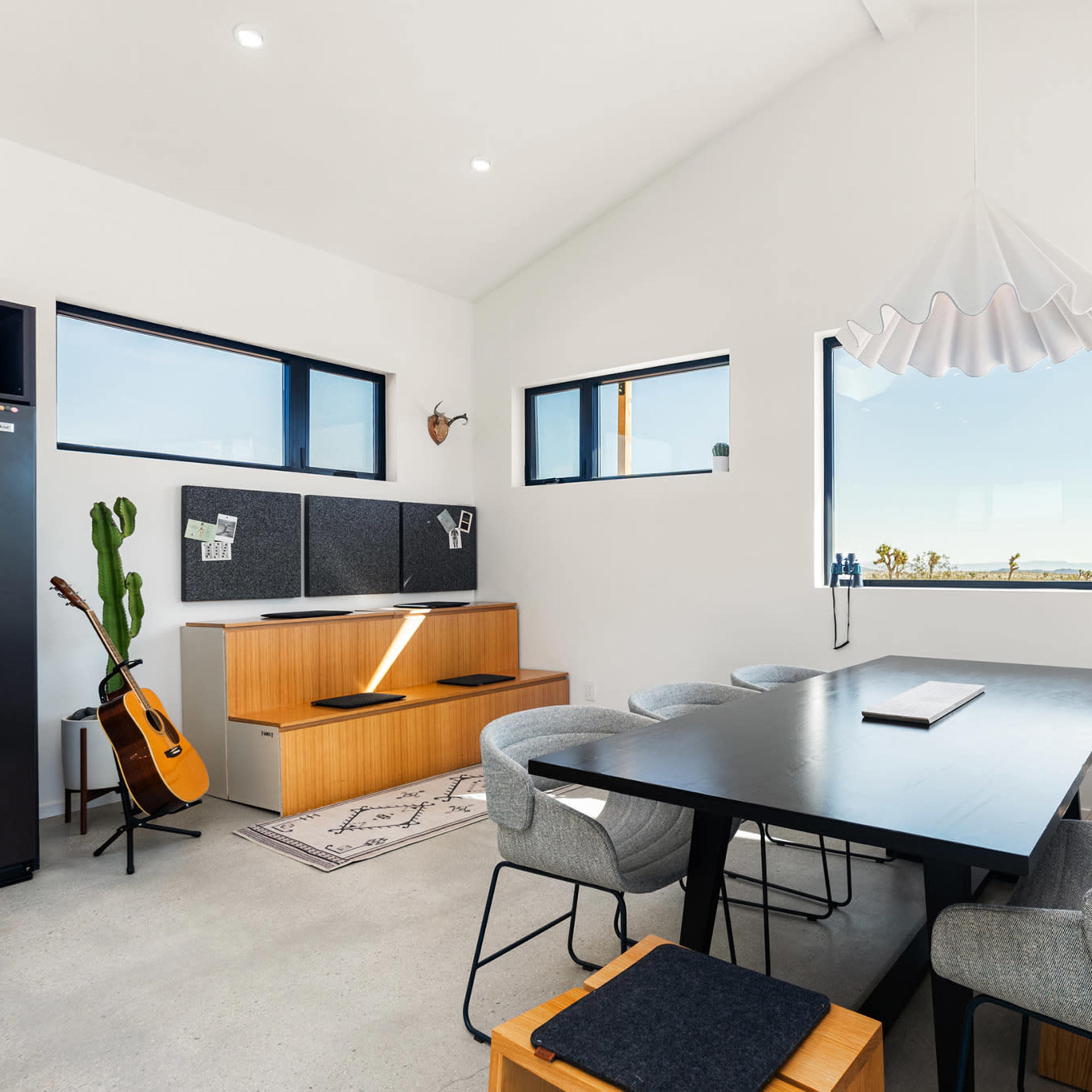 A modern dining area features a black table, gray chairs, a guitar, and large windows with a view.