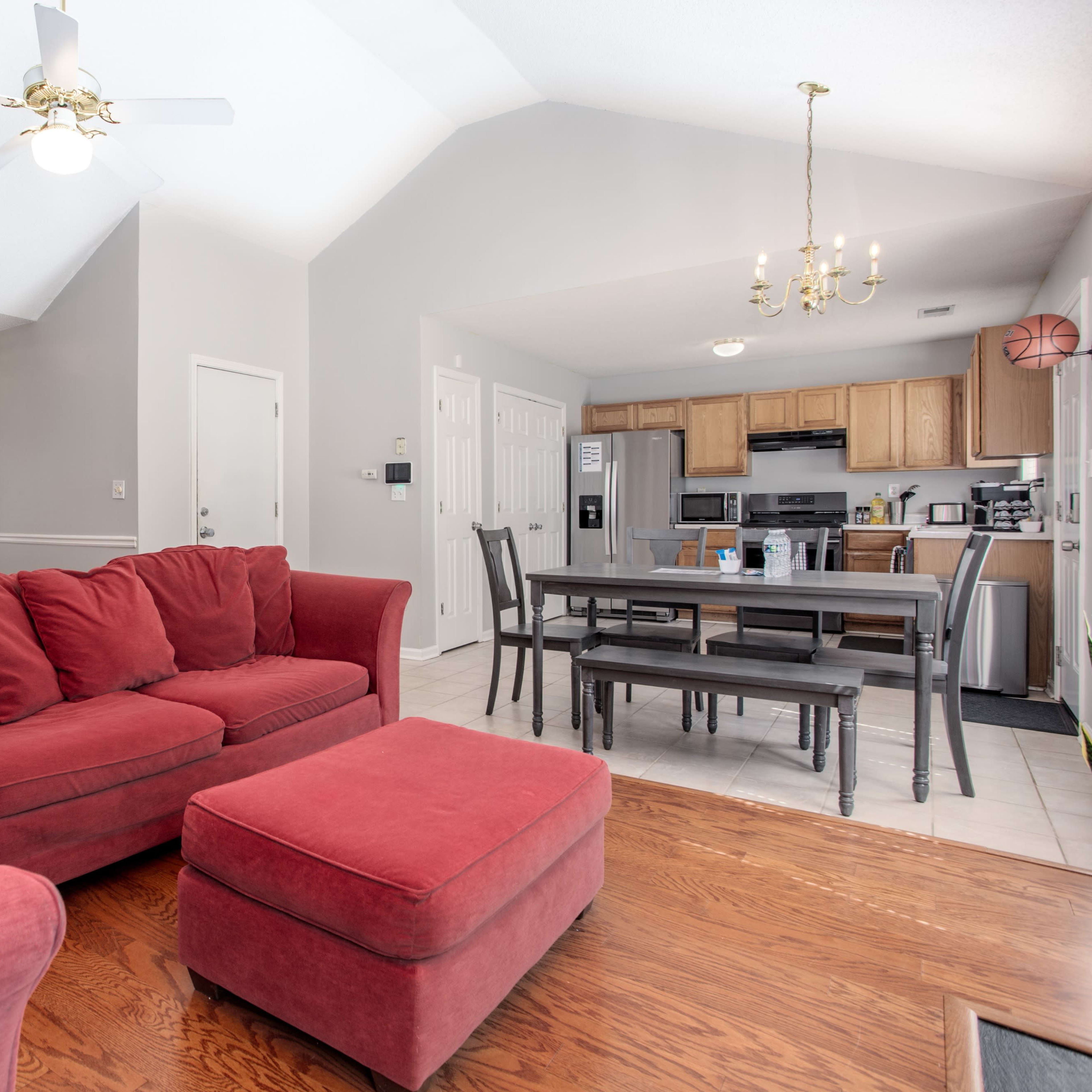A living area features a red couch and ottoman, a dining table, and an open kitchen with wooden cabinets.
