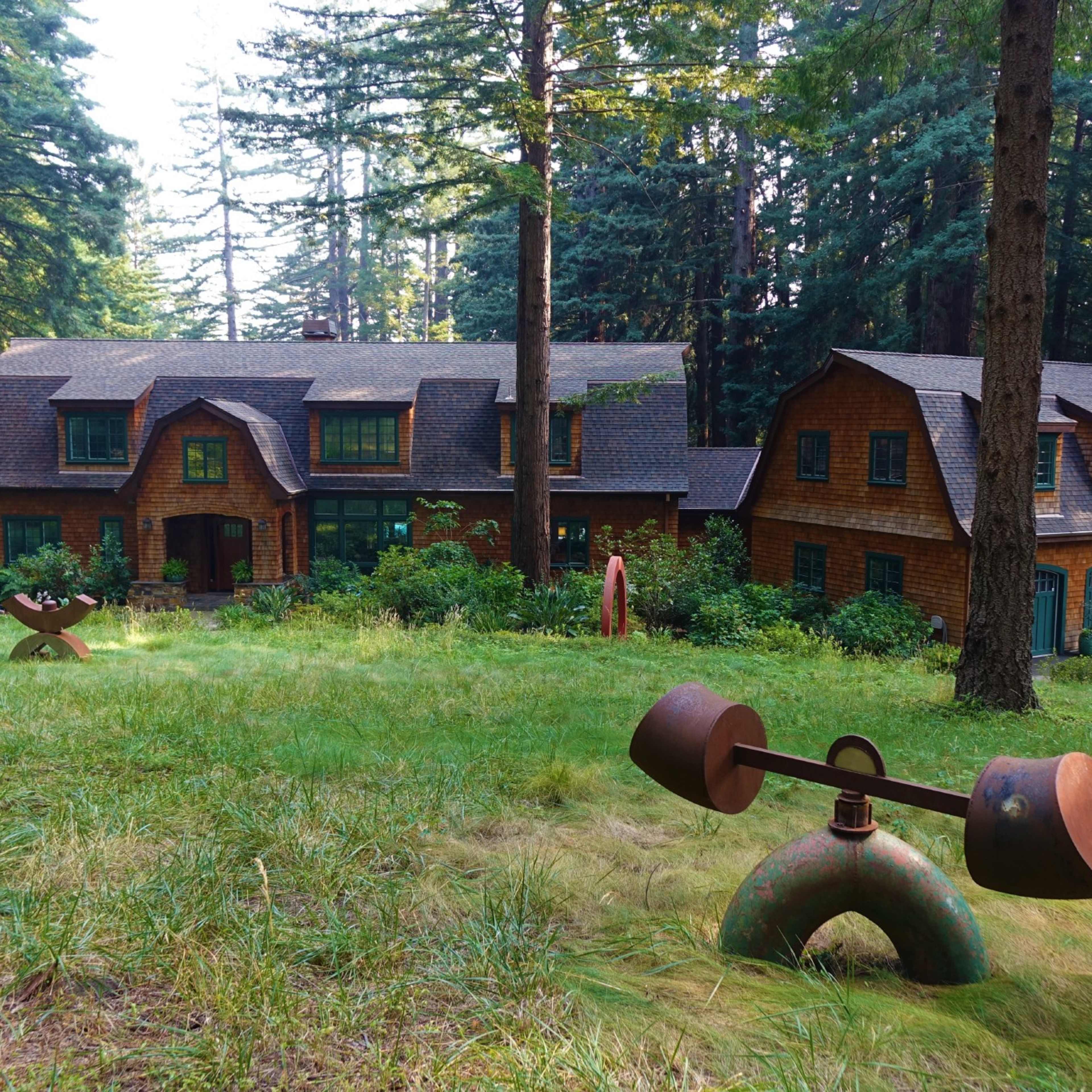 A large wooden house with multiple gabled roofs is surrounded by tall trees and lawn sculptures in a wooded area.