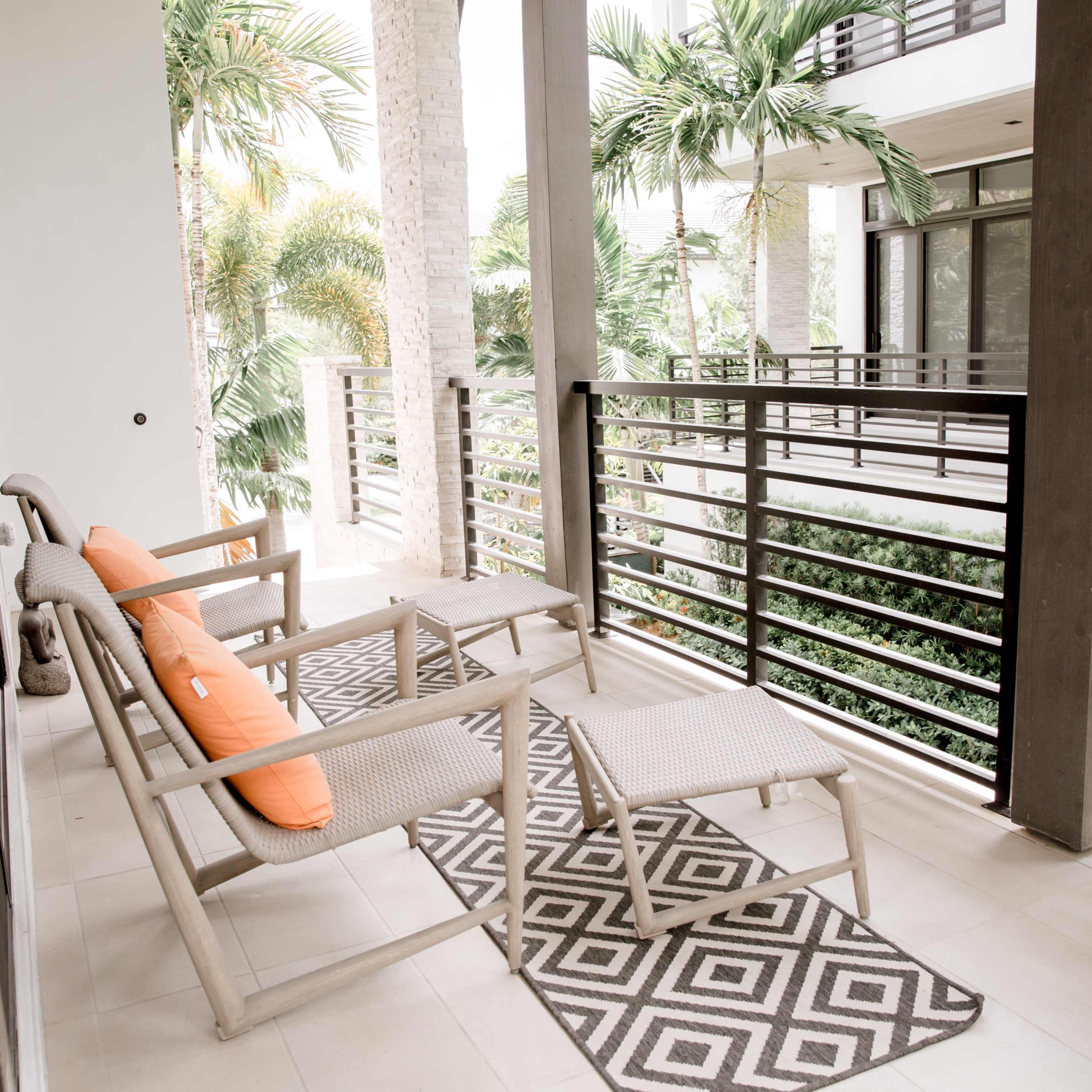 A balcony with two lounge chairs, an orange cushion, and a geometric-patterned rug surrounded by tropical plants.