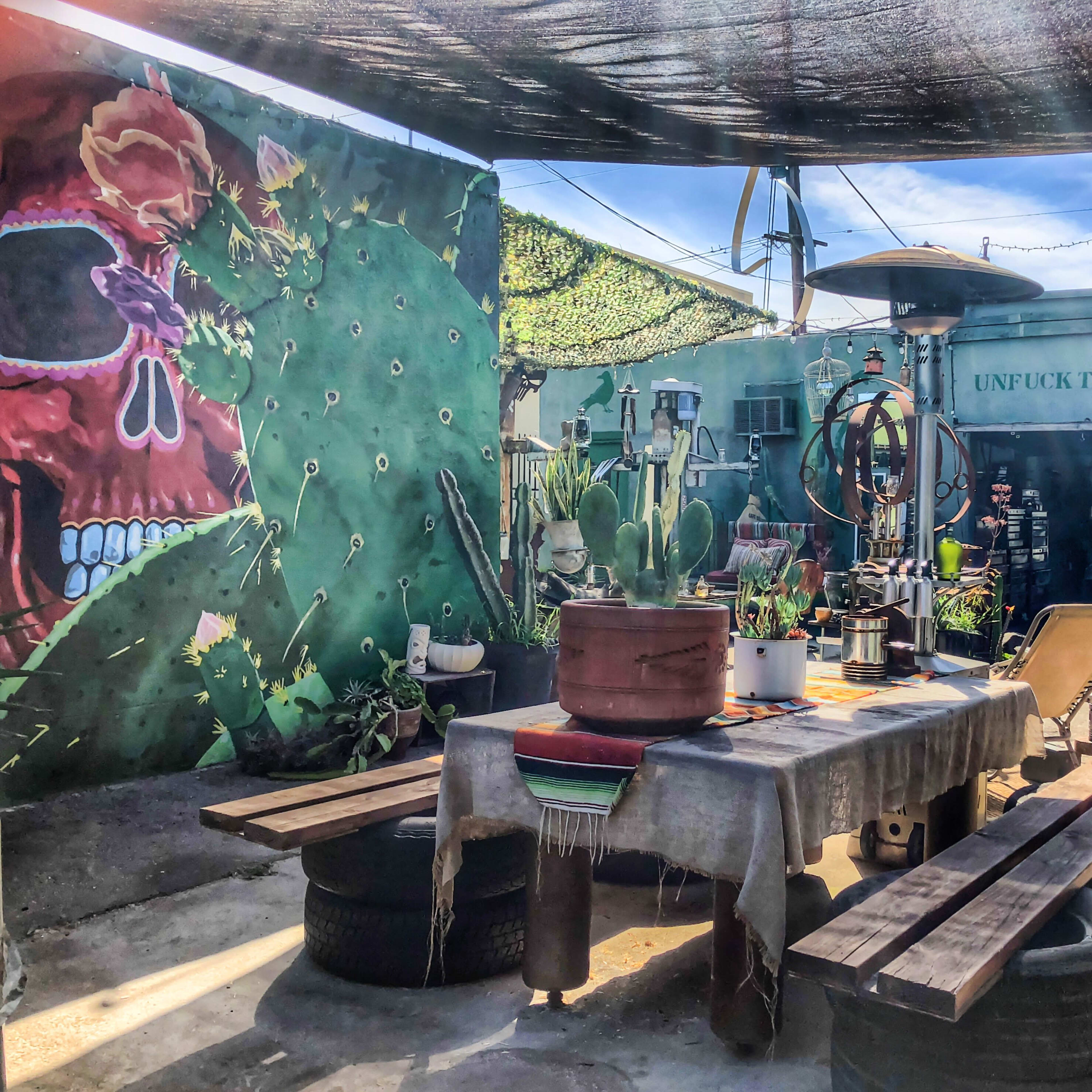 The scene shows a vibrant outdoor space featuring a wooden table surrounded by tire benches, with a mural on the wall and various plants and art pieces scattered throughout.
