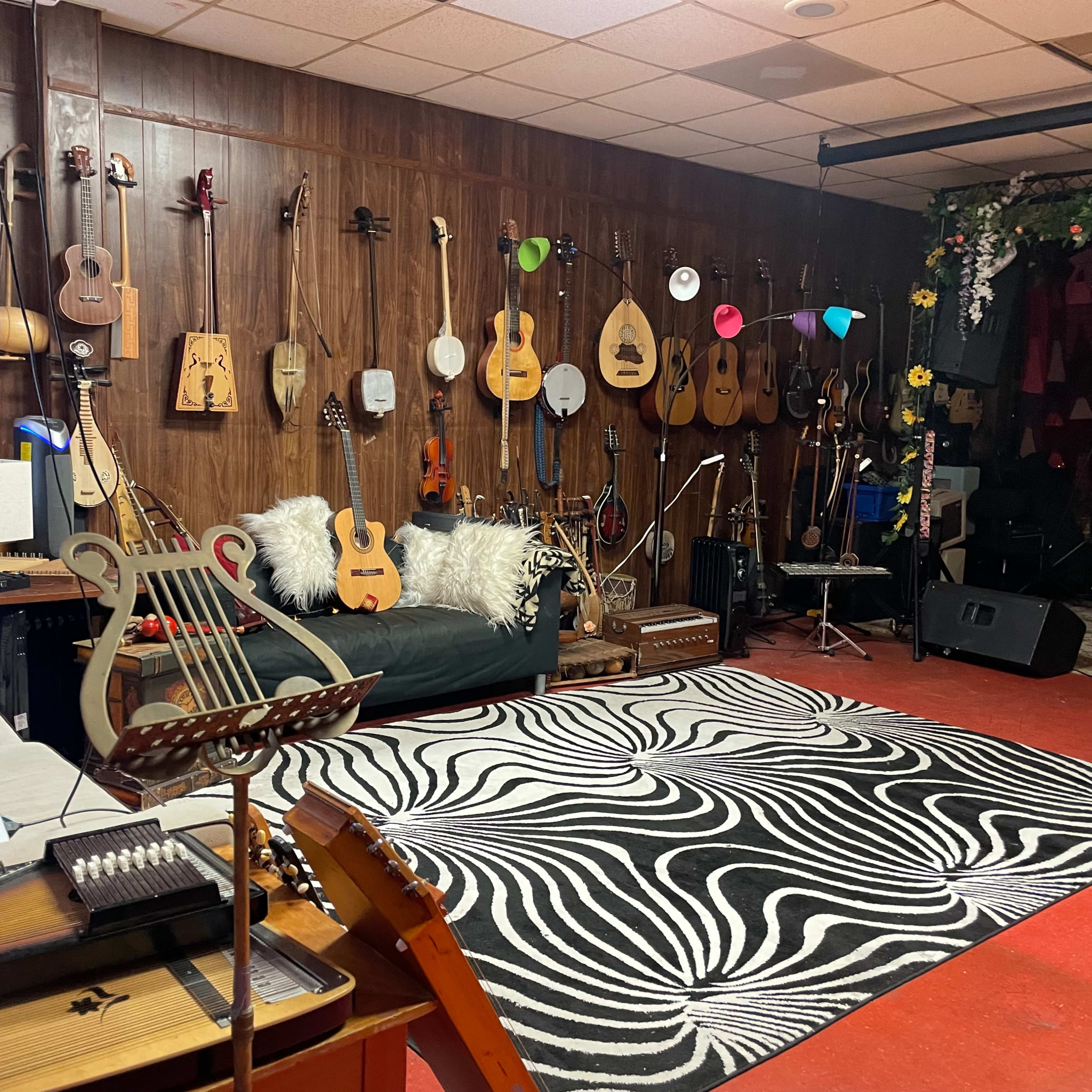 The room features a variety of string instruments hanging on a wooden wall, with a black and white patterned rug on the floor, a couch, and a small stage area for performances.