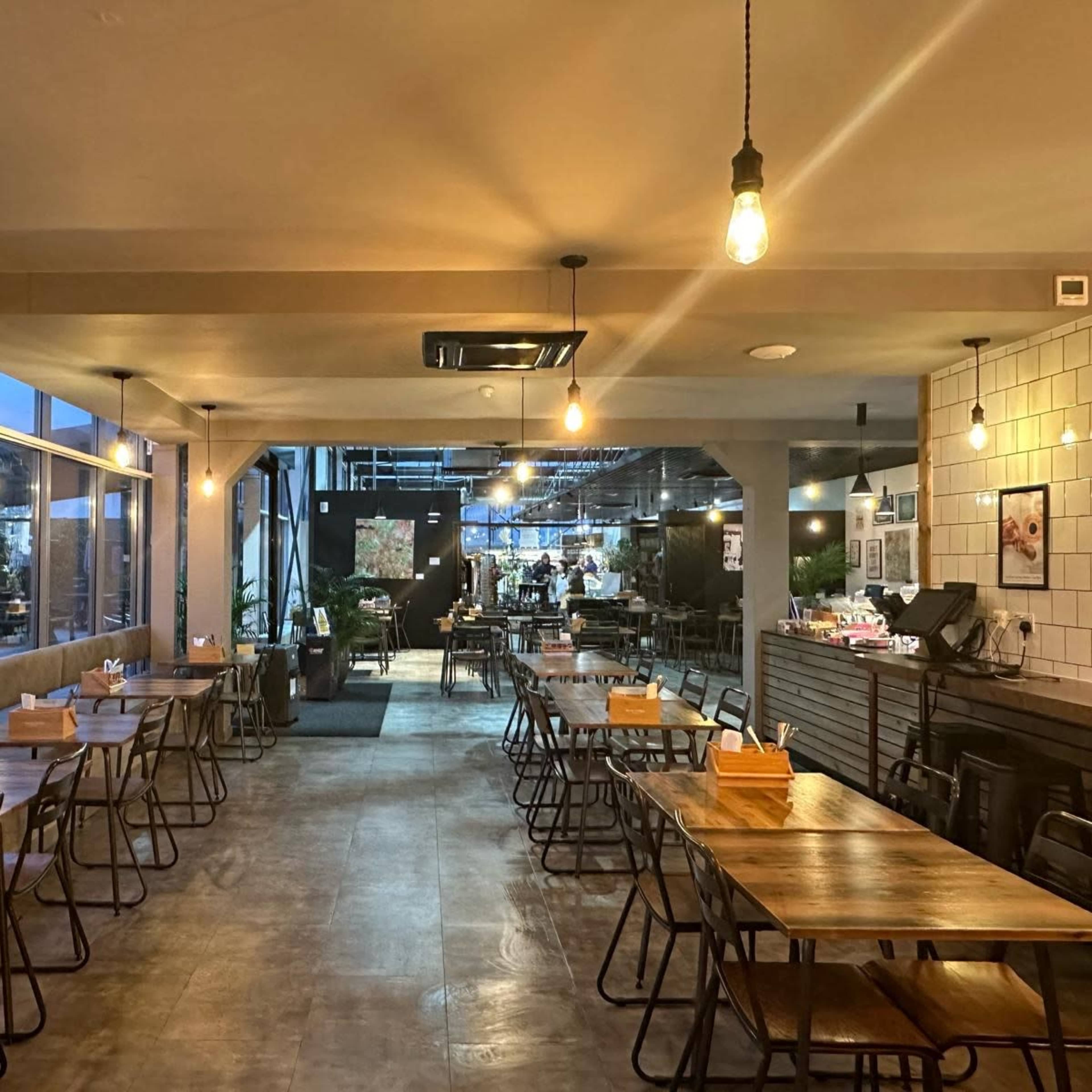 A spacious restaurant interior with wooden tables and chairs arranged in a neat configuration, illuminated by hanging light bulbs.