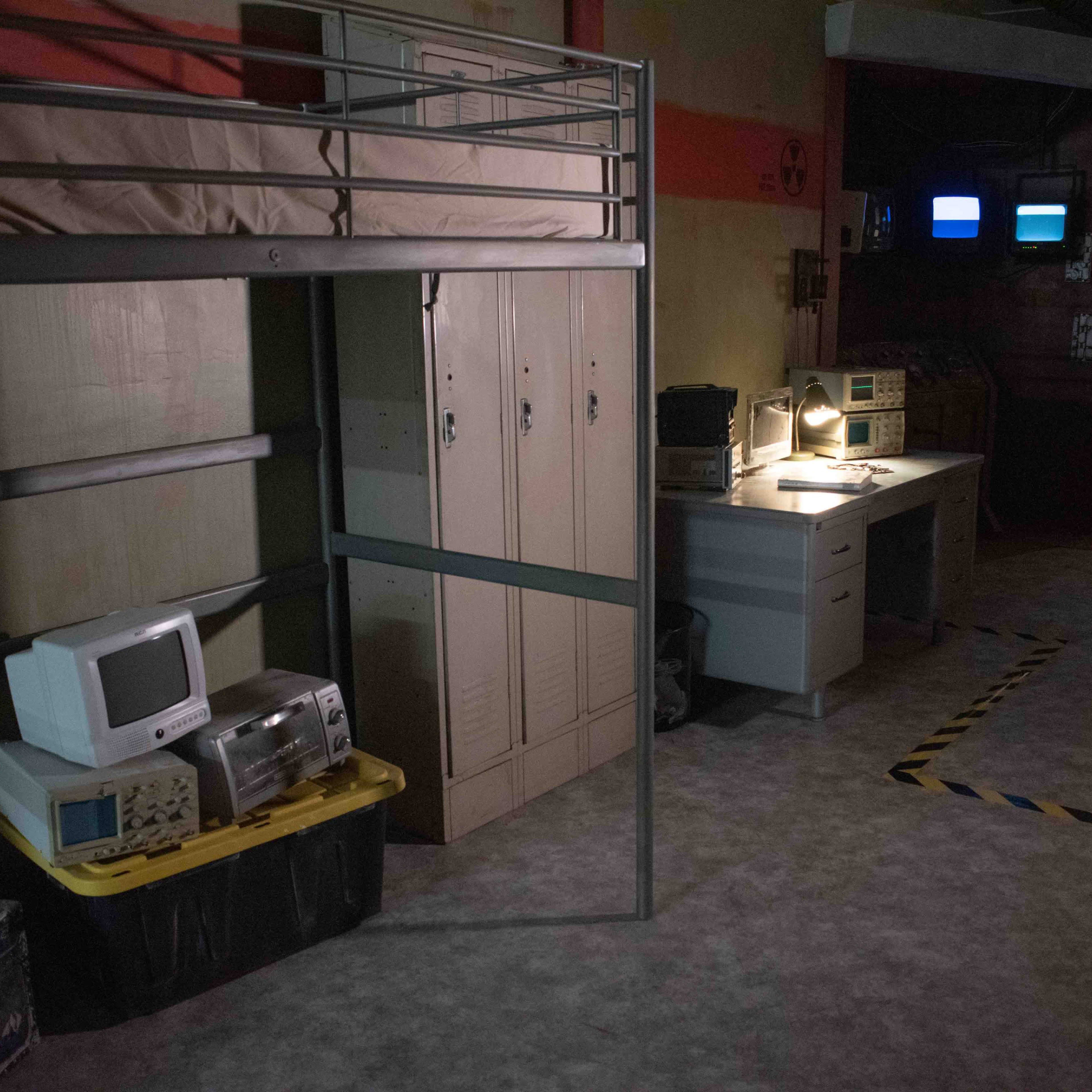 A dimly lit room with a lofted bed, lockers, an old computer, and a desk with a light and monitors.