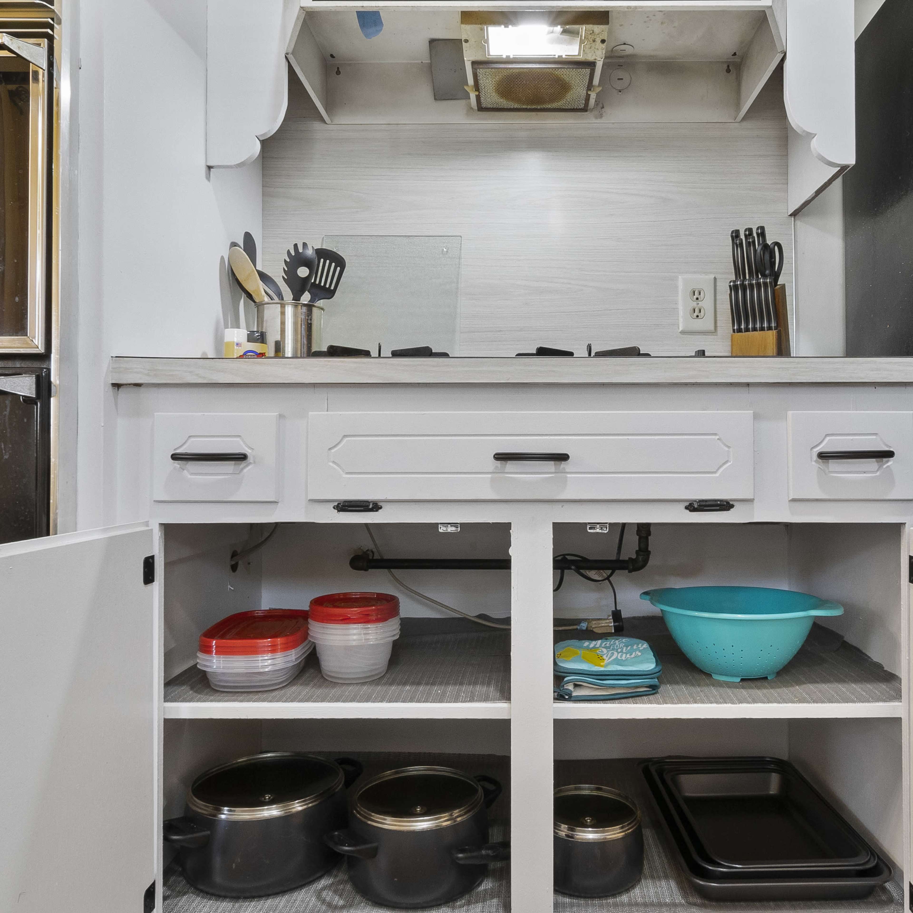A kitchen area with a stove, utensils, and storage cabinets containing pots, bowls, and a baking tray.