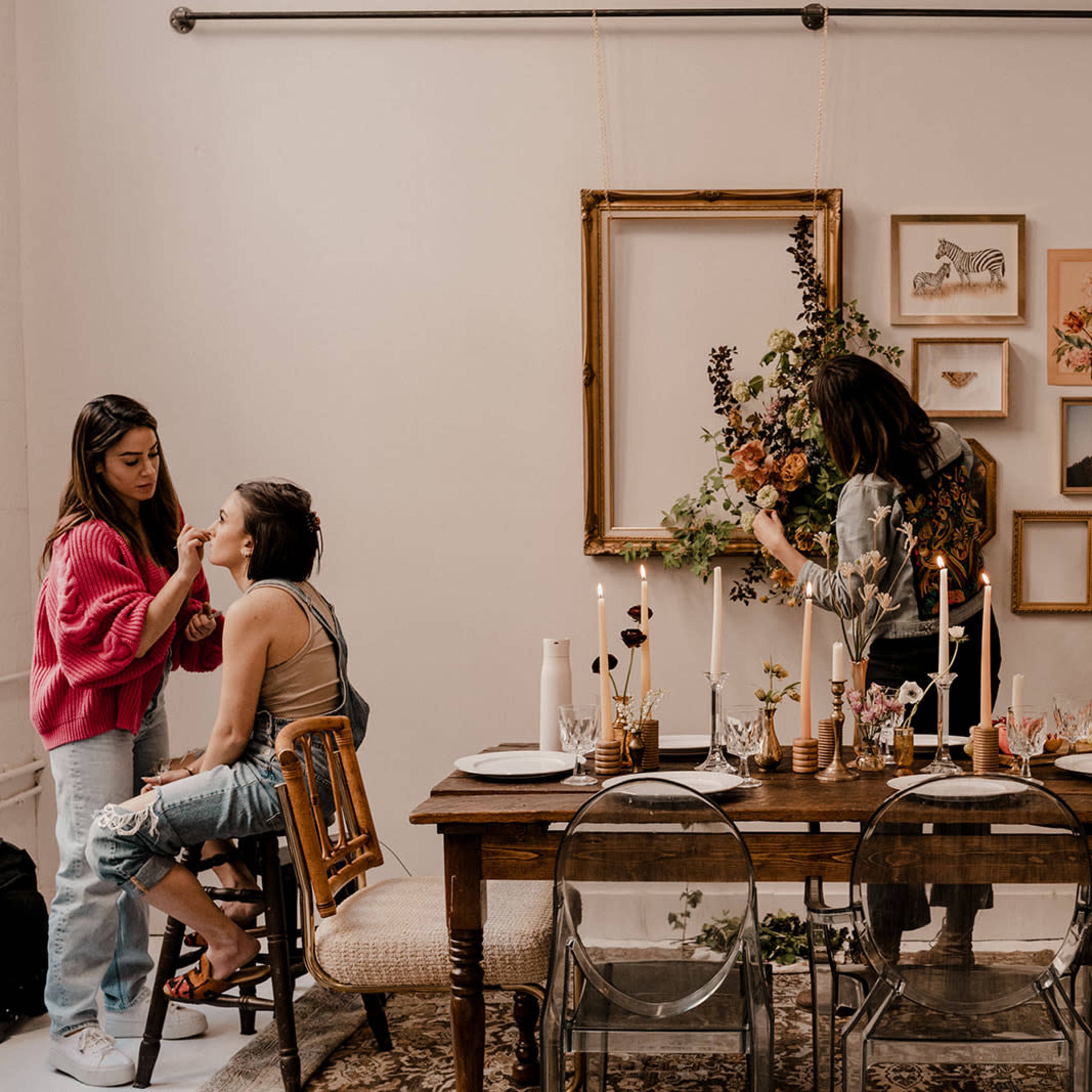A dining table is set with candles and floral decorations, while one person applies makeup to another and another arranges flowers on a wall.