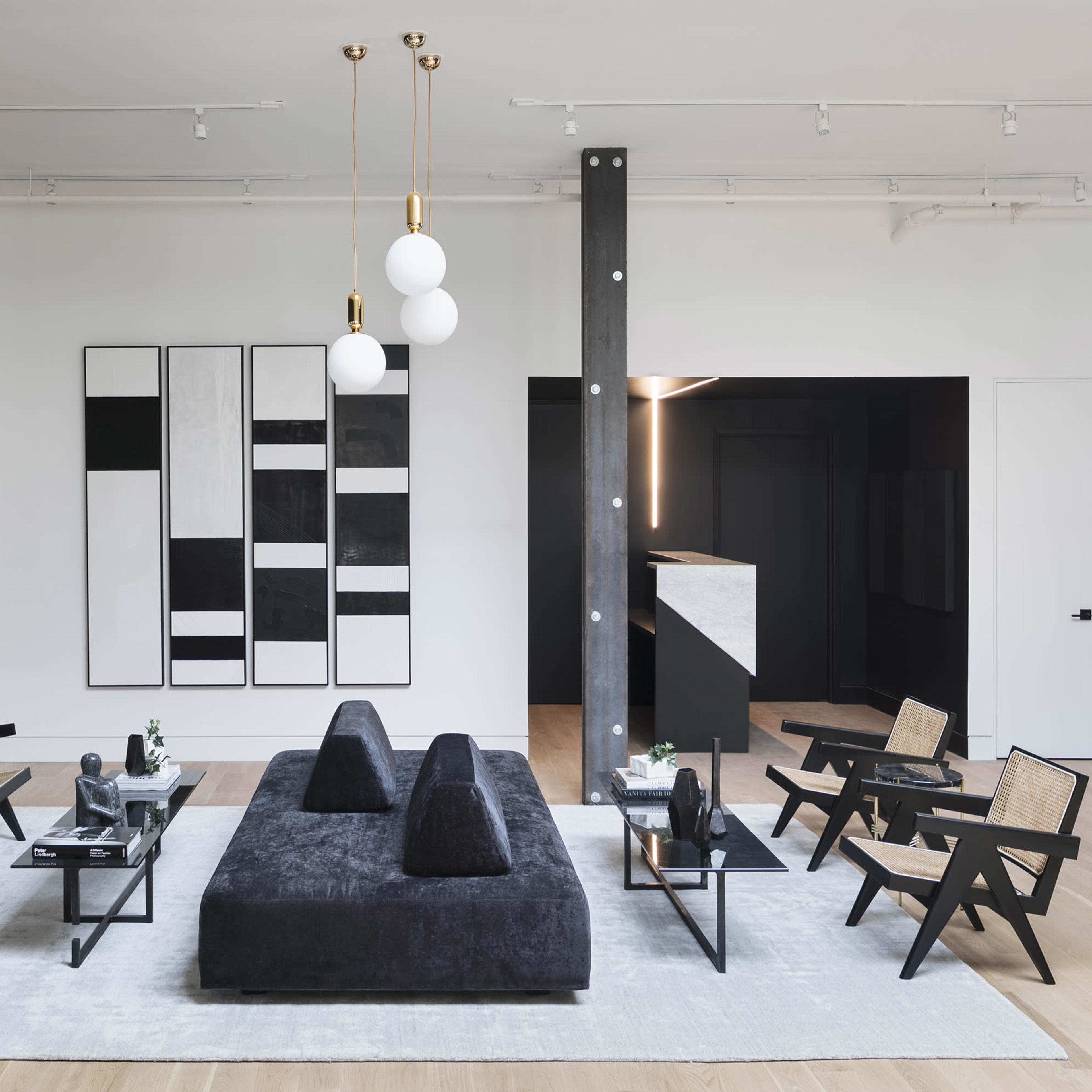 A modern living space features a central black seating arrangement surrounded by minimalist chairs, with art on the walls and large windows letting in natural light.