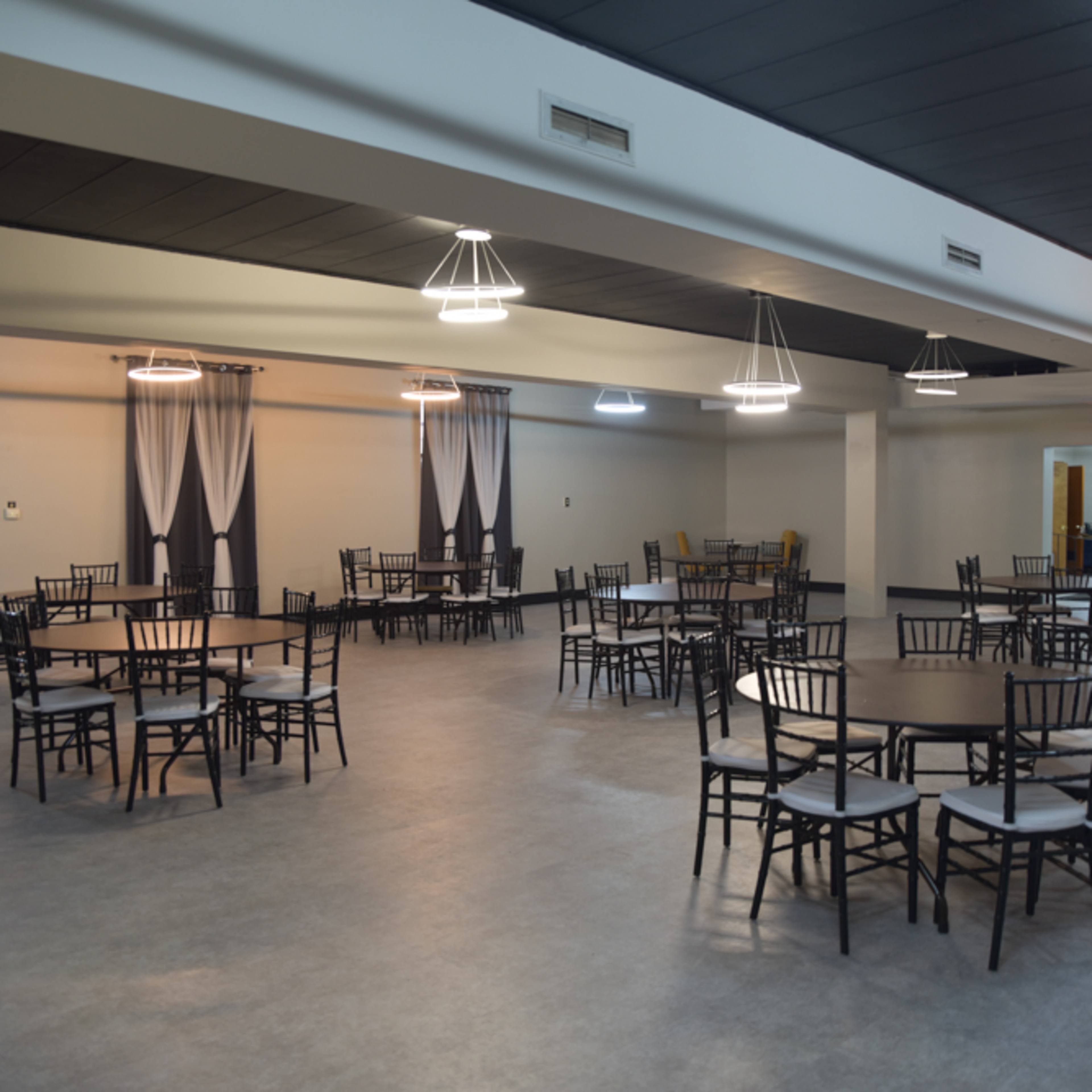 A spacious, empty dining hall with several round tables and black chairs arranged neatly under modern pendant lights.