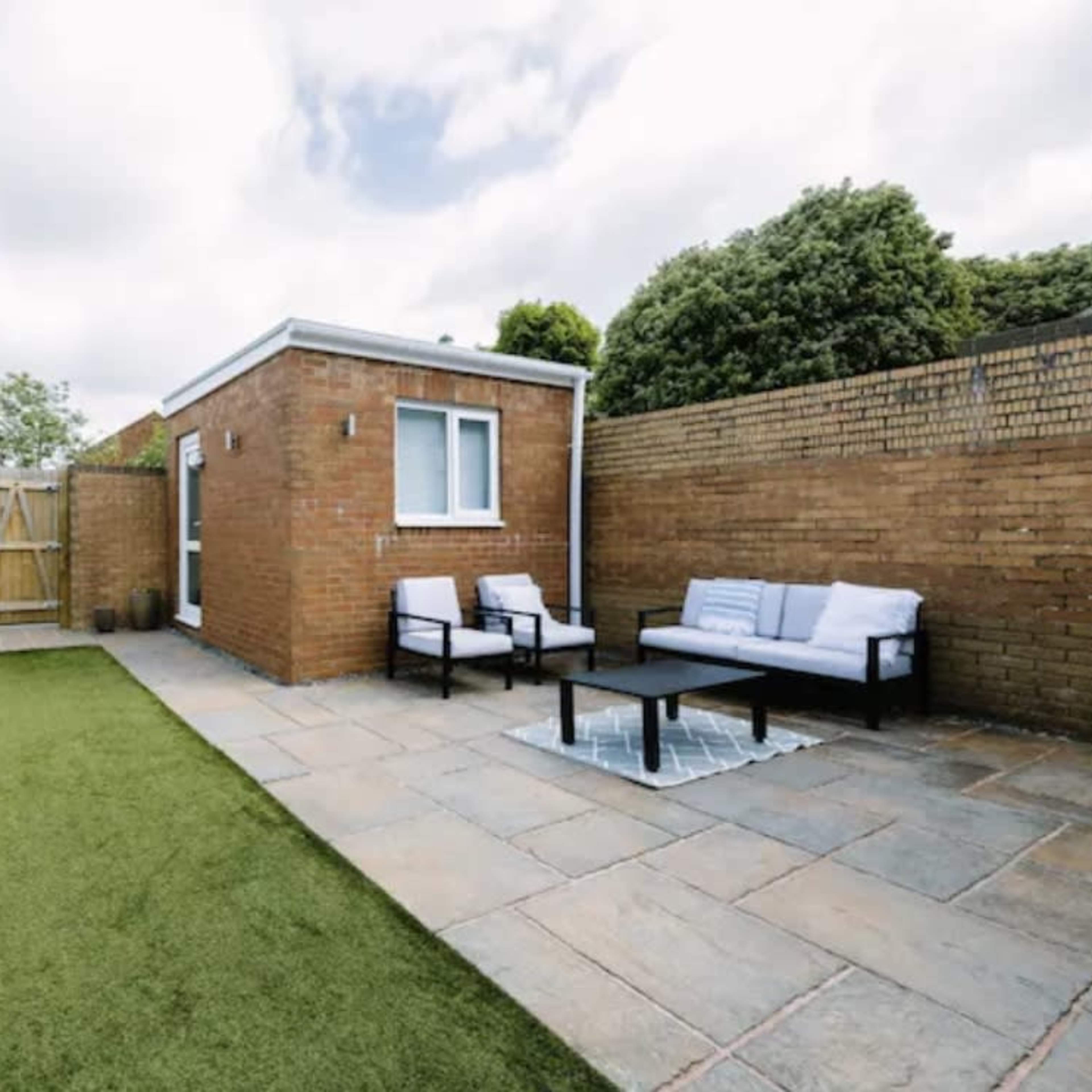 A small garden area featuring a brick structure, a paved patio with a table and chairs, and artificial grass surrounded by wooden fencing.