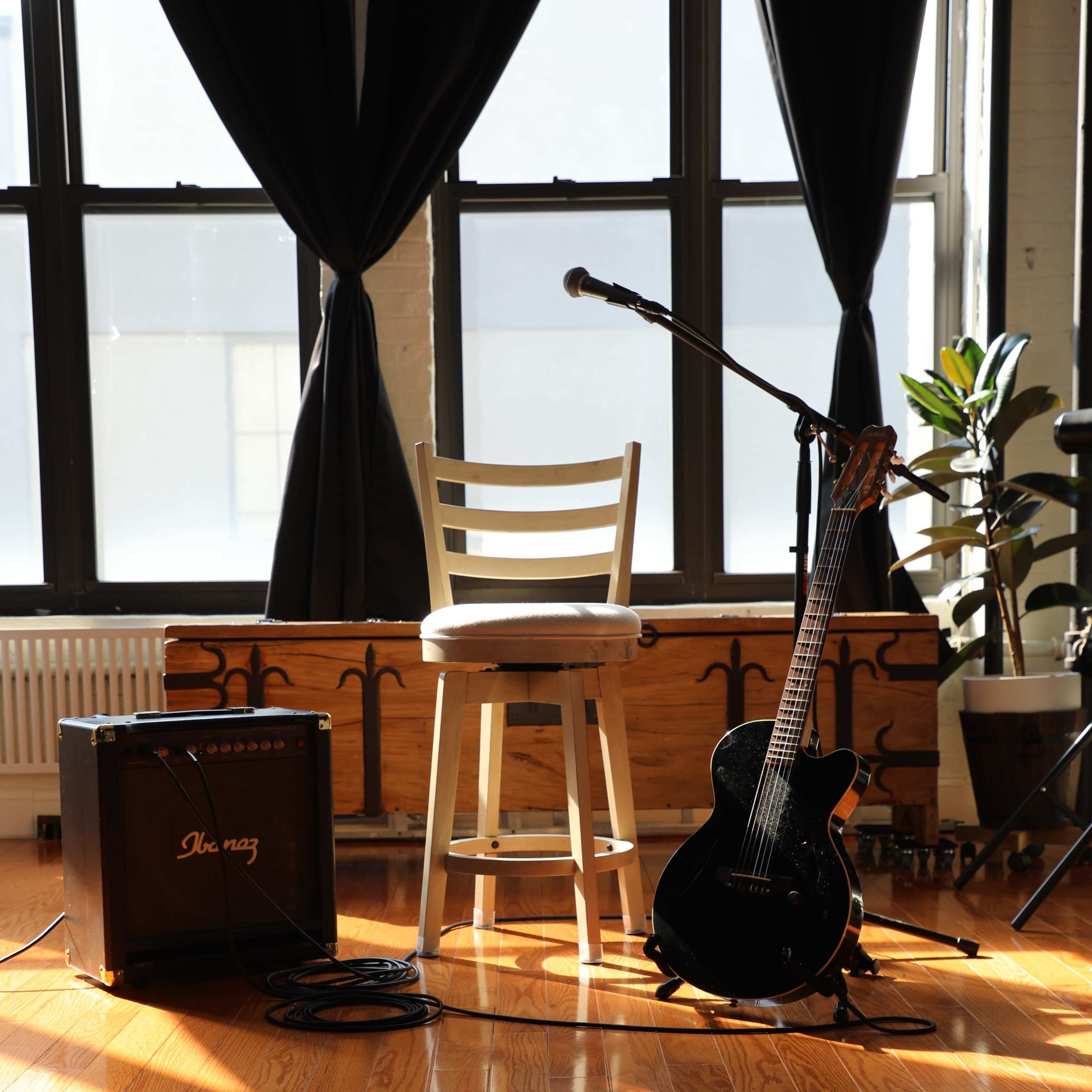 The scene features a stool next to a microphone, an electric guitar, and an amplifier, all set on a shiny wooden floor with large windows in the background.