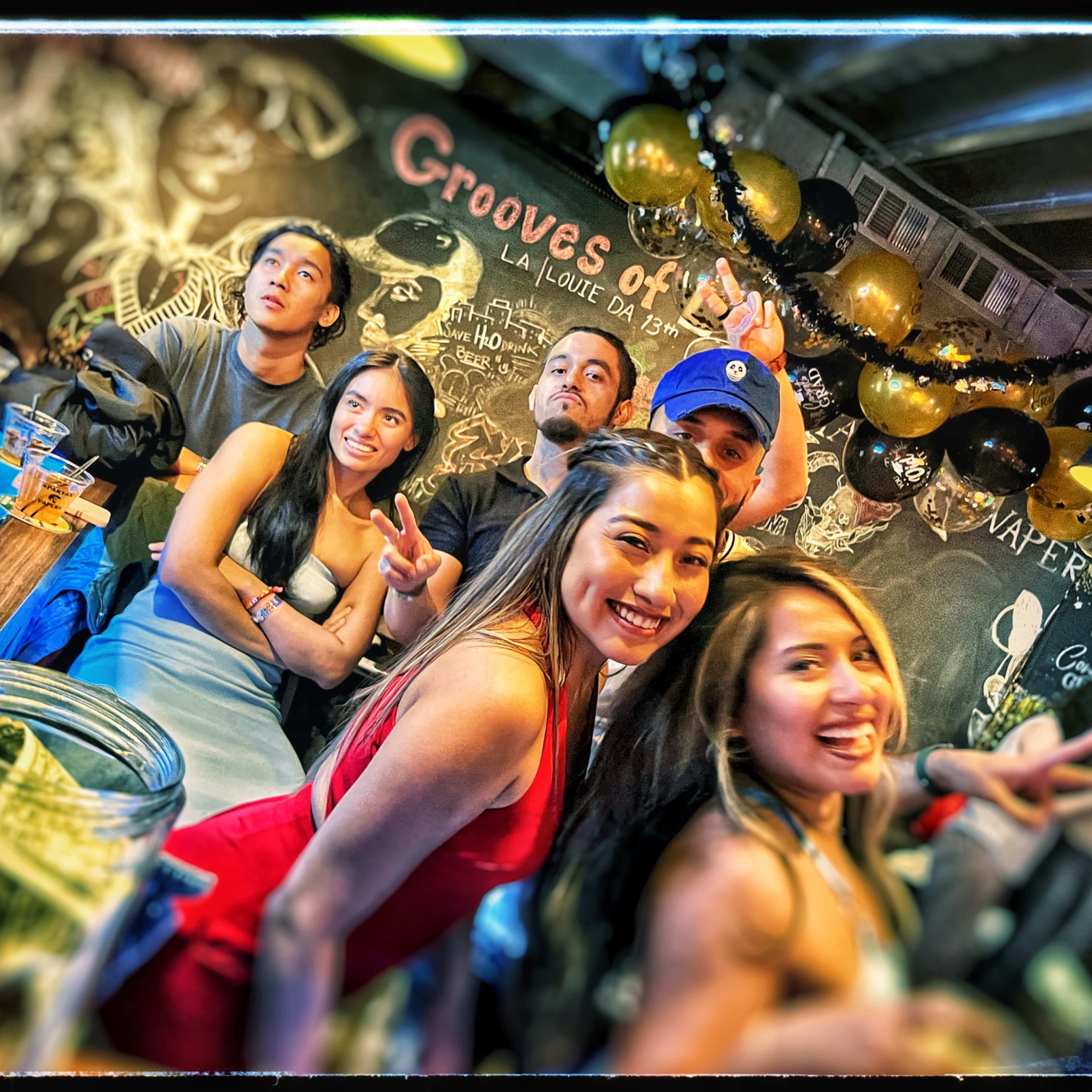 A group of six people poses playfully at a bar decorated with balloons and art on the black wall behind them.