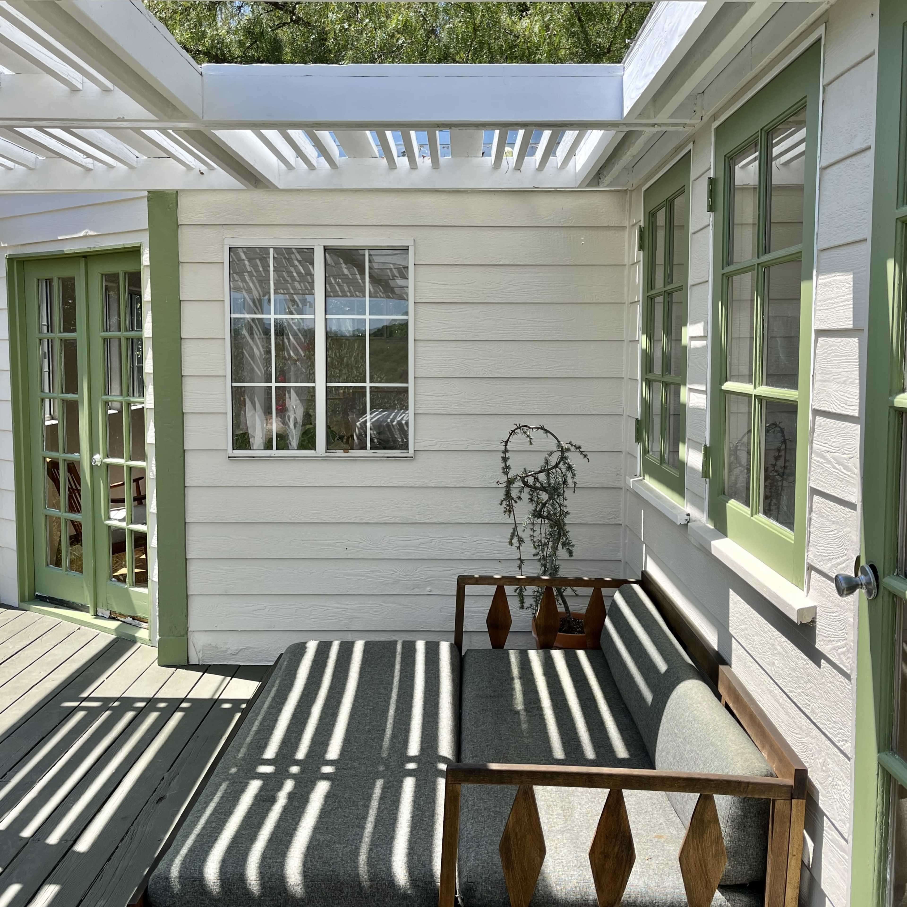 A daylight scene shows a wooden deck with a lounging space featuring a gray cushion, surrounded by green-trimmed windows and white walls under a slatted roof.