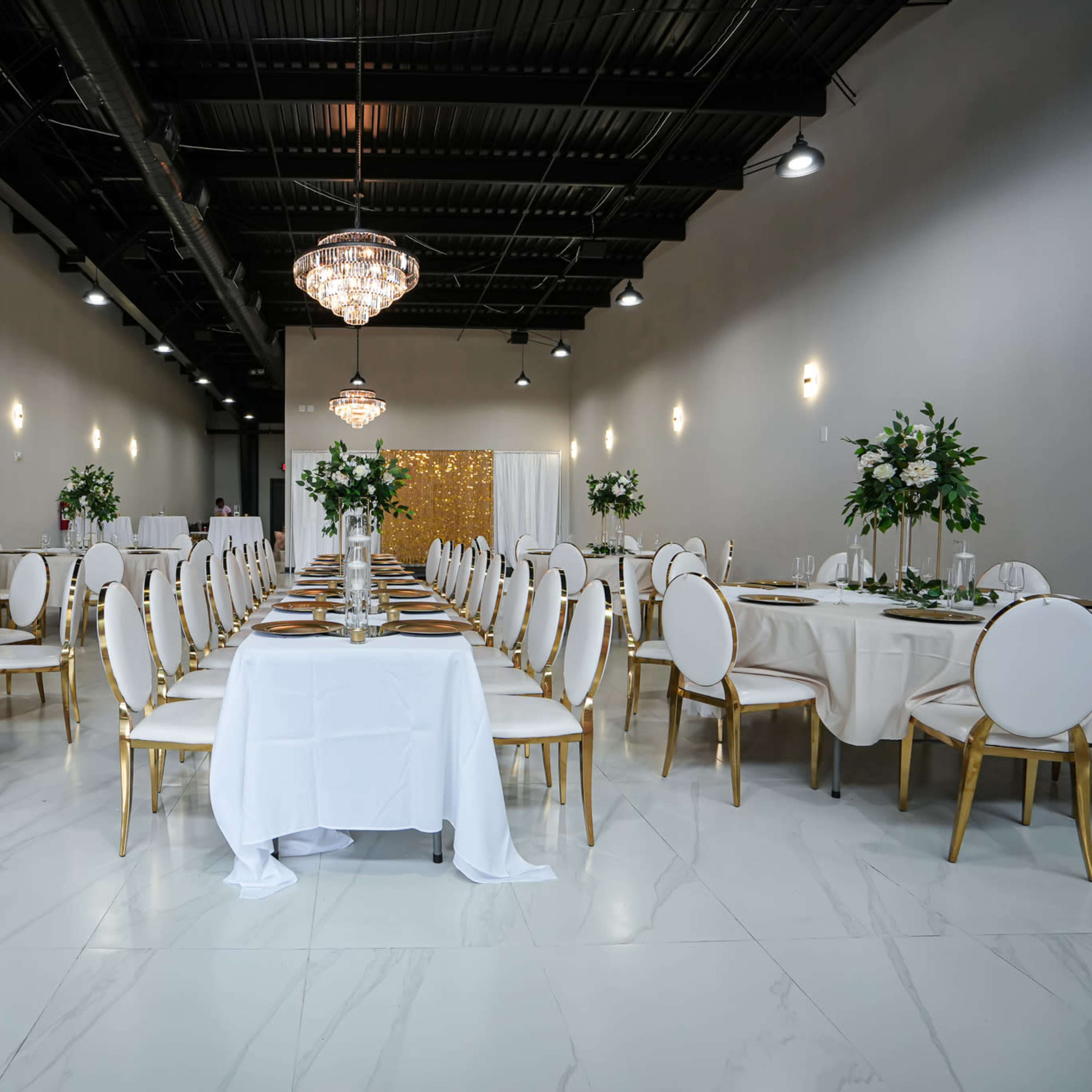 A spacious banquet hall is set up with round tables decorated with white tablecloths and gold-accented chairs, featuring centerpieces of greenery and crystal chandeliers overhead.