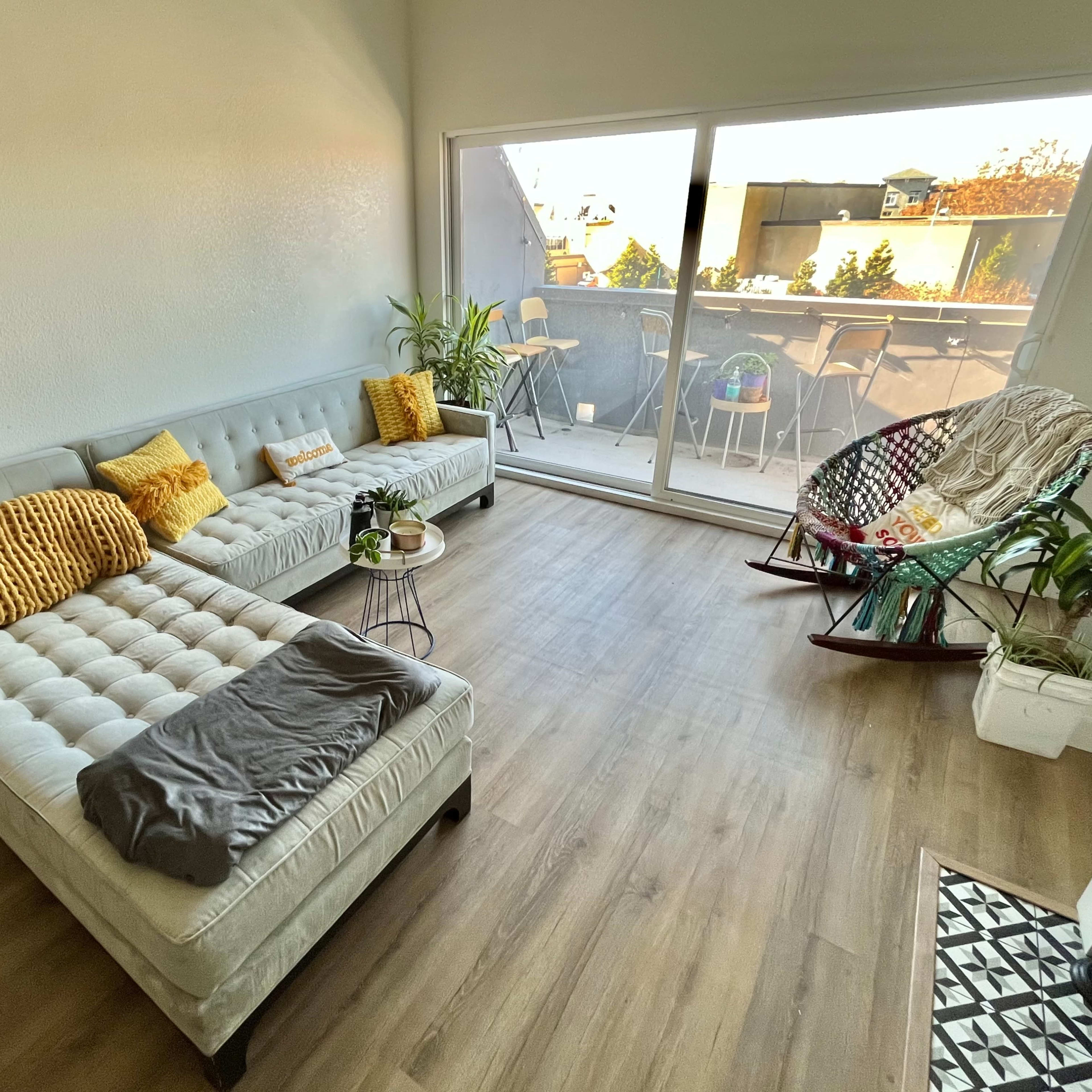 The room features a light-filled space with a gray sectional sofa, a rocking chair, a small table, and plants, leading to a balcony with outdoor seating.