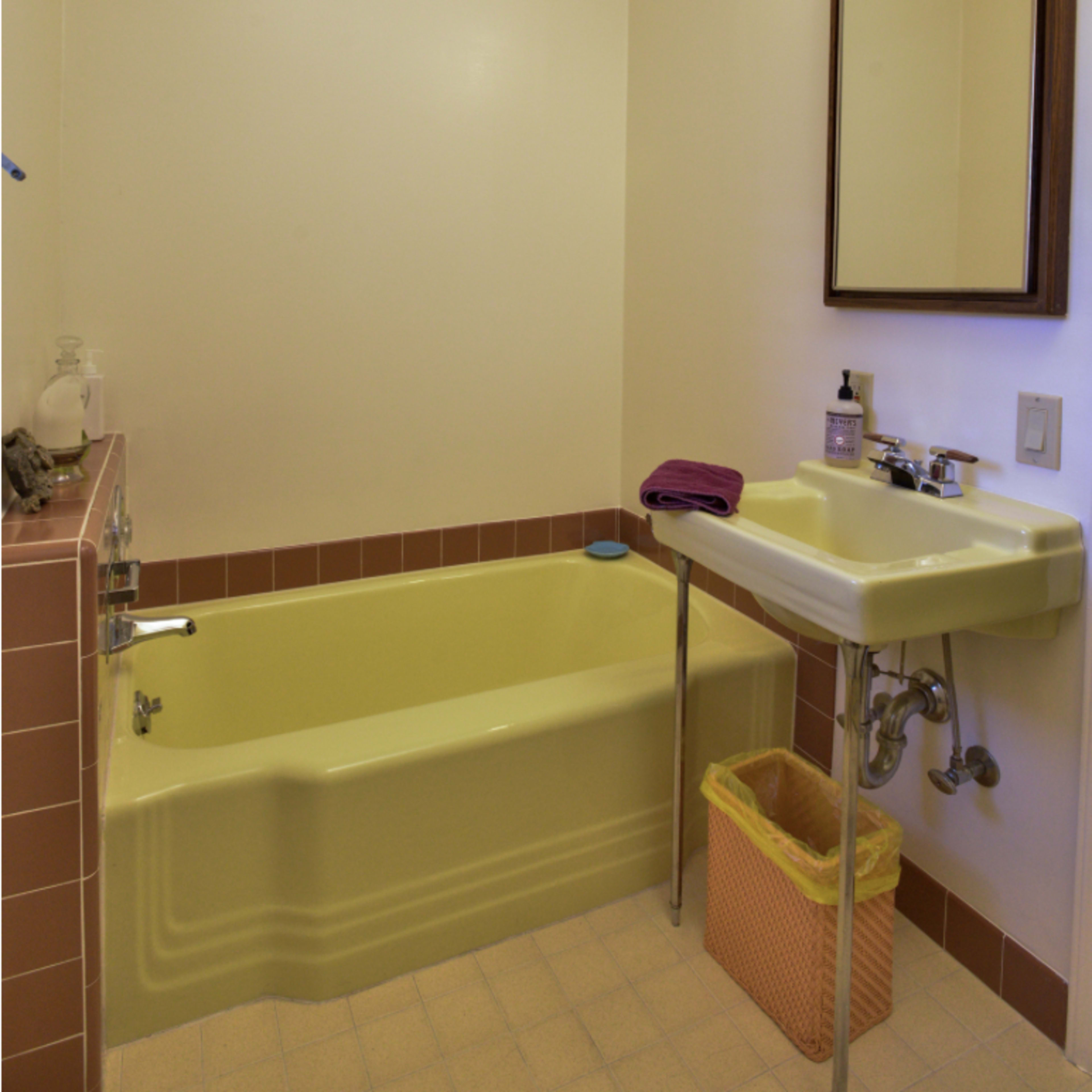 A bathroom featuring a yellow bathtub, a sink with exposed plumbing, and a mirror above the sink, along with pink tile accents.