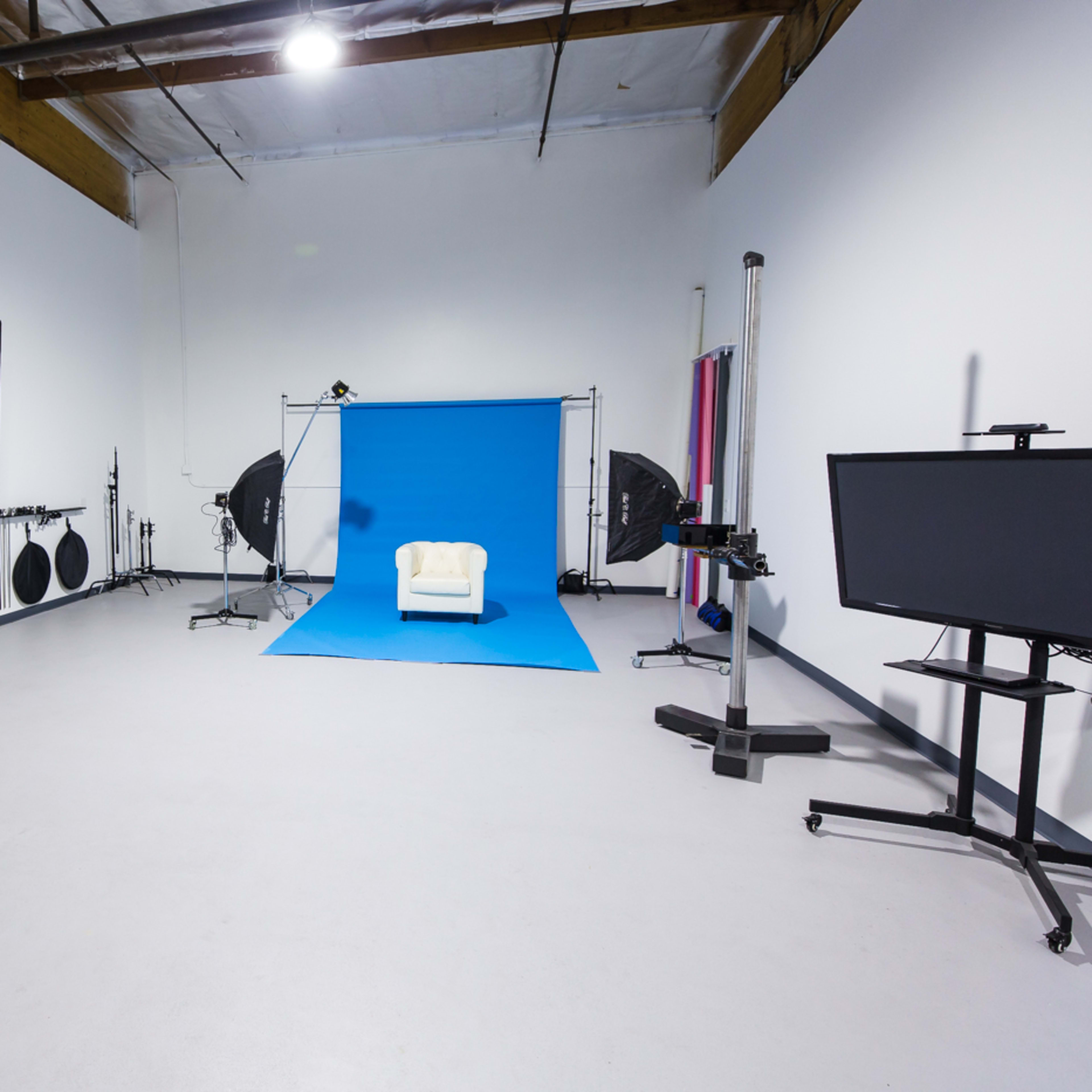 A photography studio features a white chair on a blue backdrop, with various lighting equipment and a monitor positioned around the space.