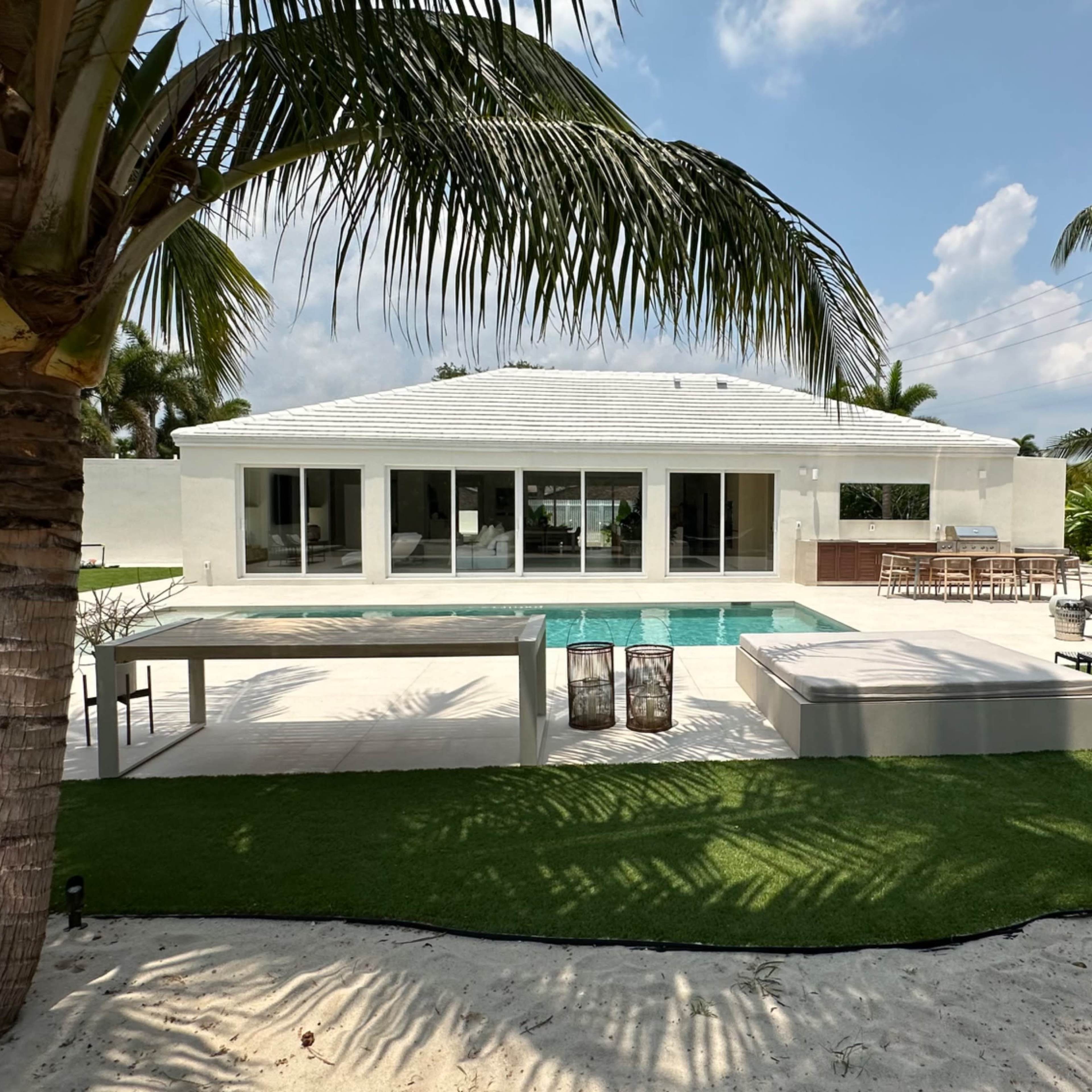 A modern house with large glass windows, a swimming pool, and a landscaped outdoor area featuring palm trees and lounge furniture.