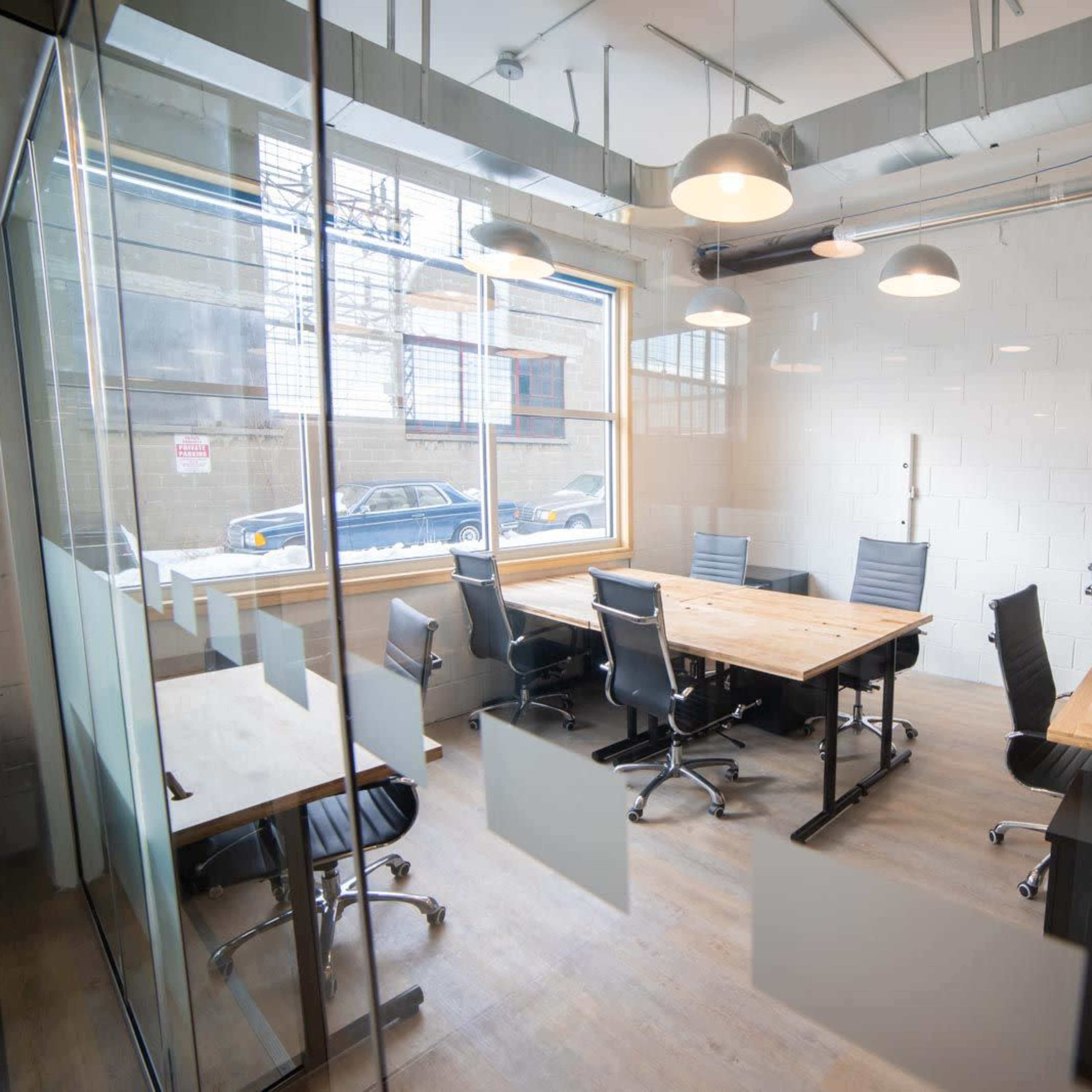 A conference room features a wooden table surrounded by black chairs and large windows, allowing natural light to enter.