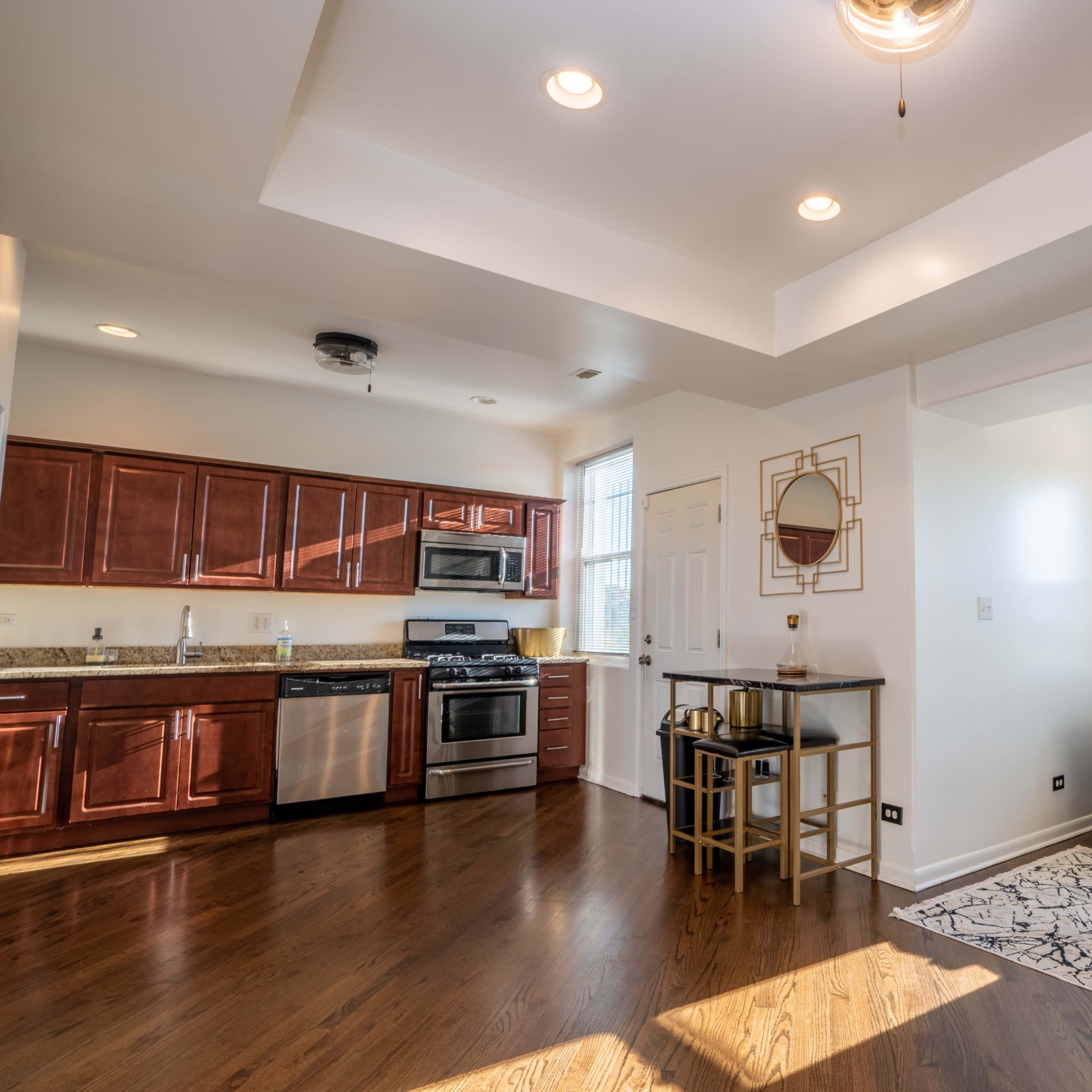 A modern kitchen and living area with wooden cabinetry, stainless steel appliances, and a gray sofa facing a coffee table on a patterned rug.