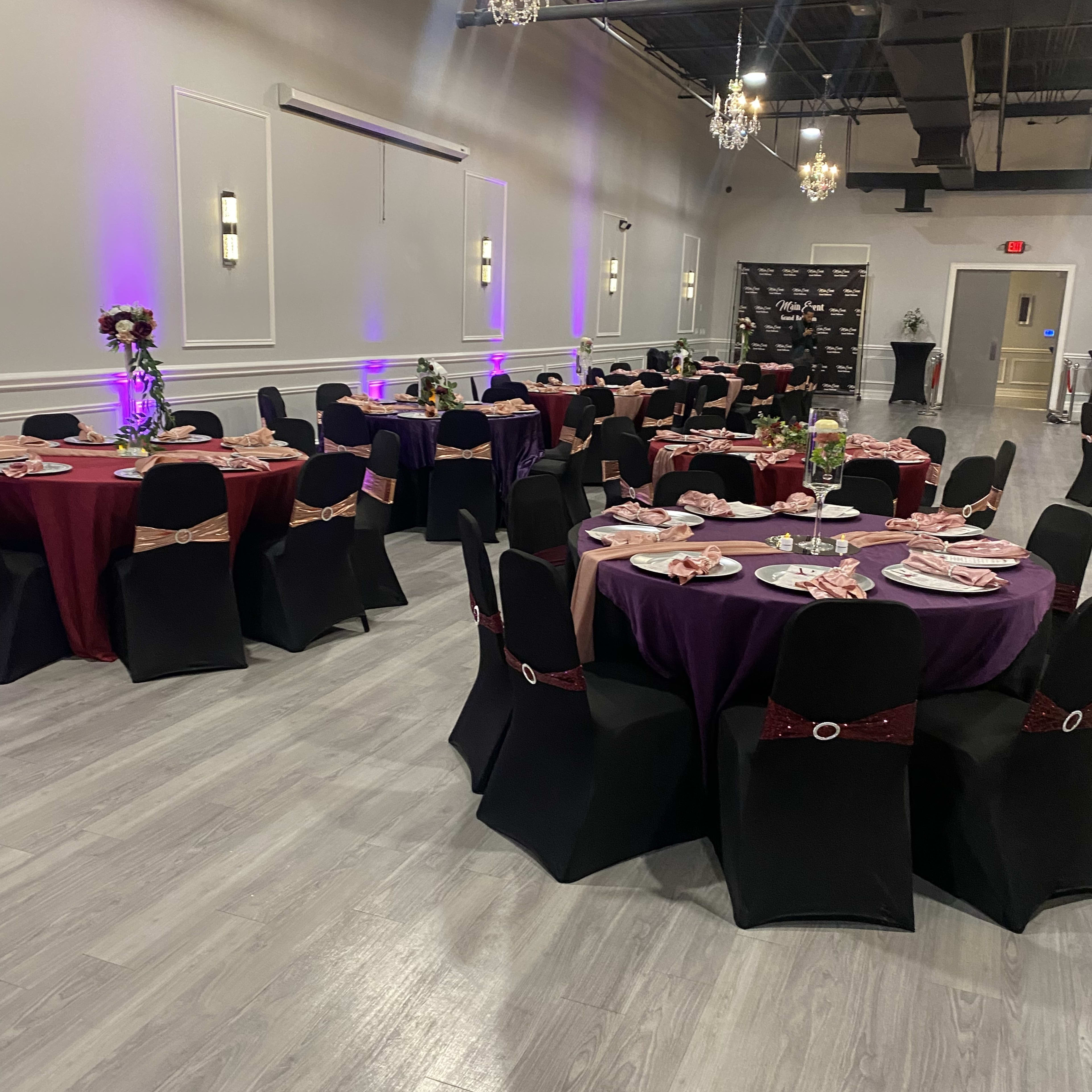 A banquet hall set up with round tables covered in black and burgundy tablecloths, adorned with centerpieces and place settings, under decorative chandeliers.