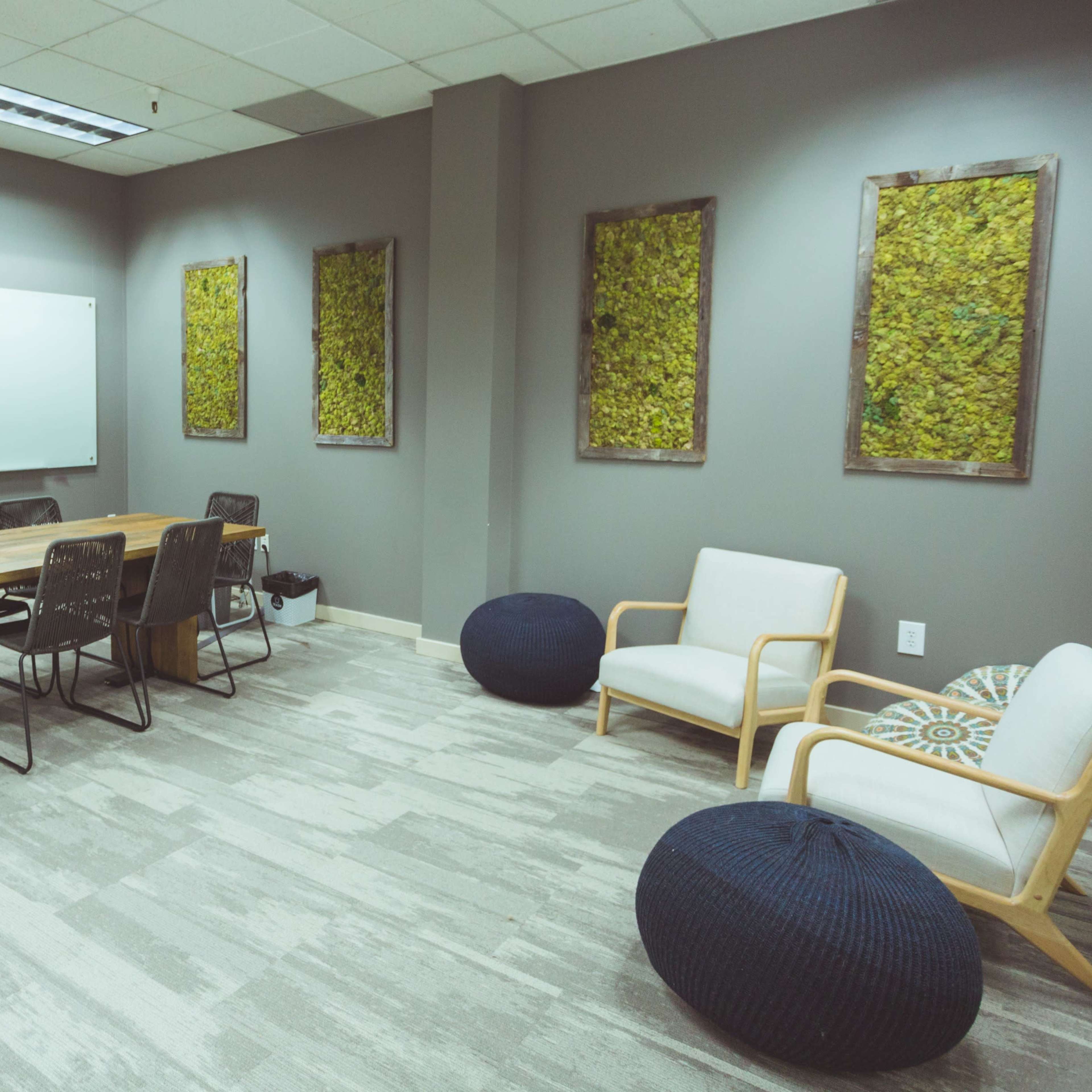 A conference room features a wooden table surrounded by chairs, with a whiteboard on one wall and moss-covered artwork on the adjacent walls.