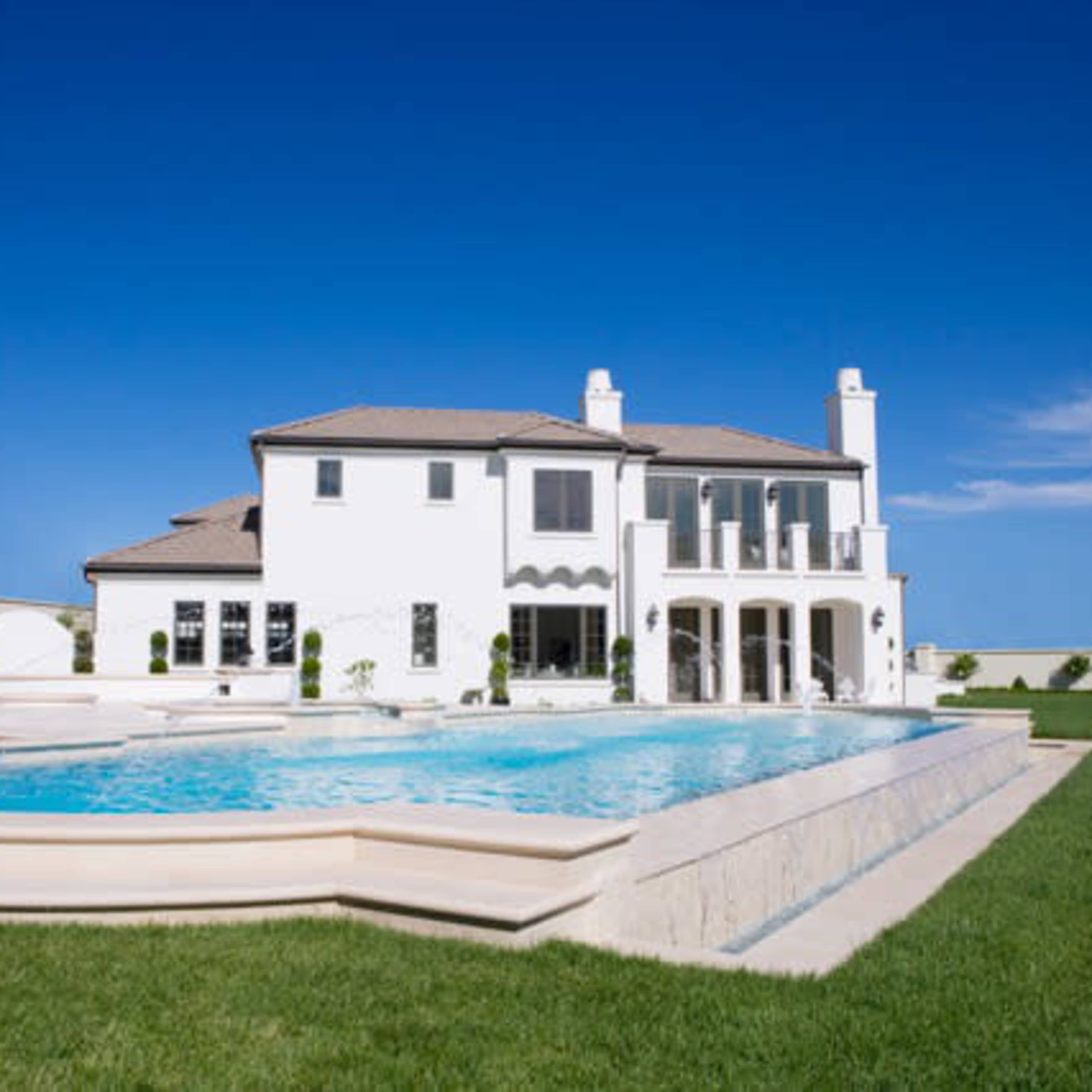 A large white house with a swimming pool in the backyard, surrounded by green grass under a clear blue sky.