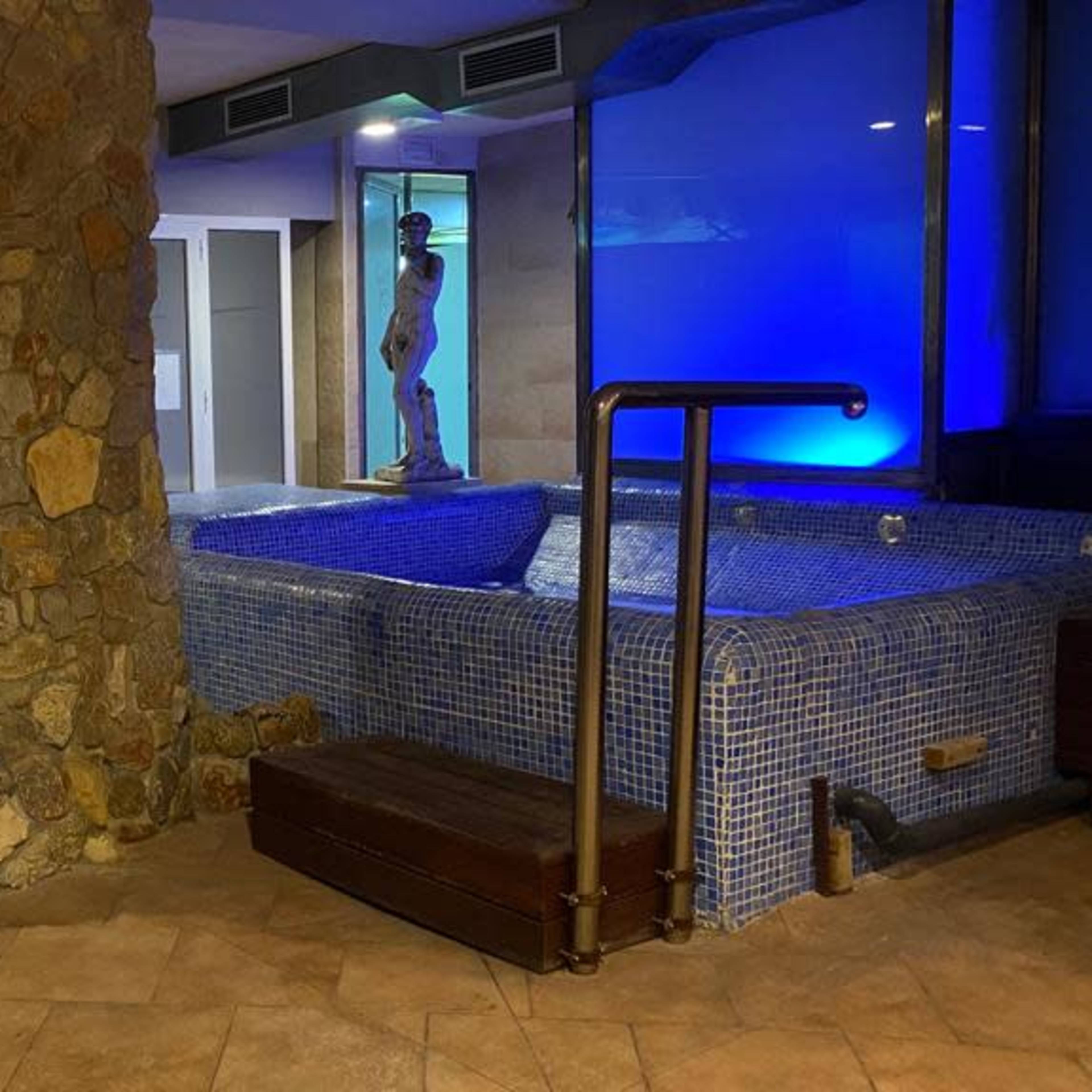 An indoor spa area featuring a hot tub, a stone pillar, a white chair, and a statue beside a blue-lit wall.