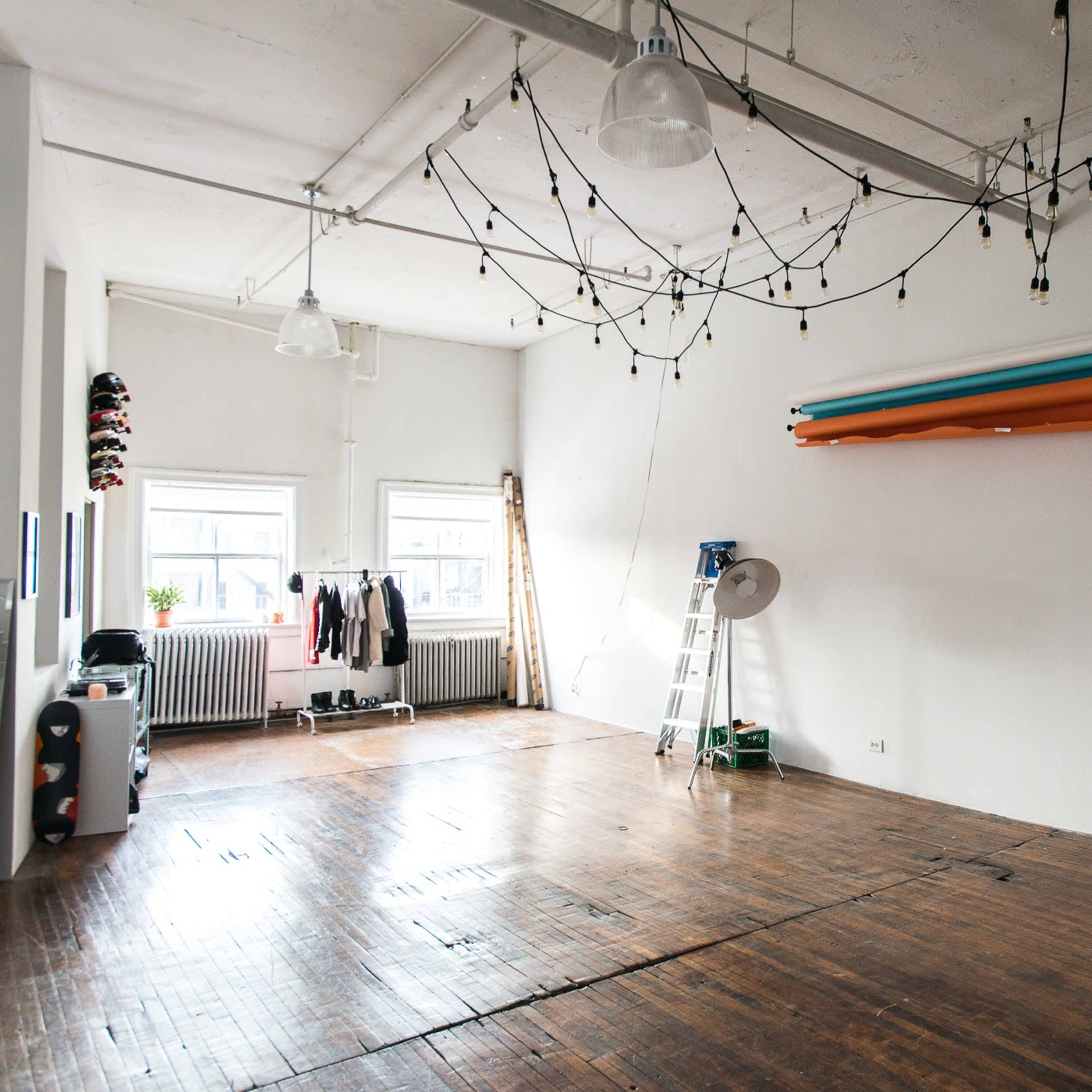 A spacious, minimally furnished photography studio with wooden flooring, a ladder, and backdrop equipment against a white wall.