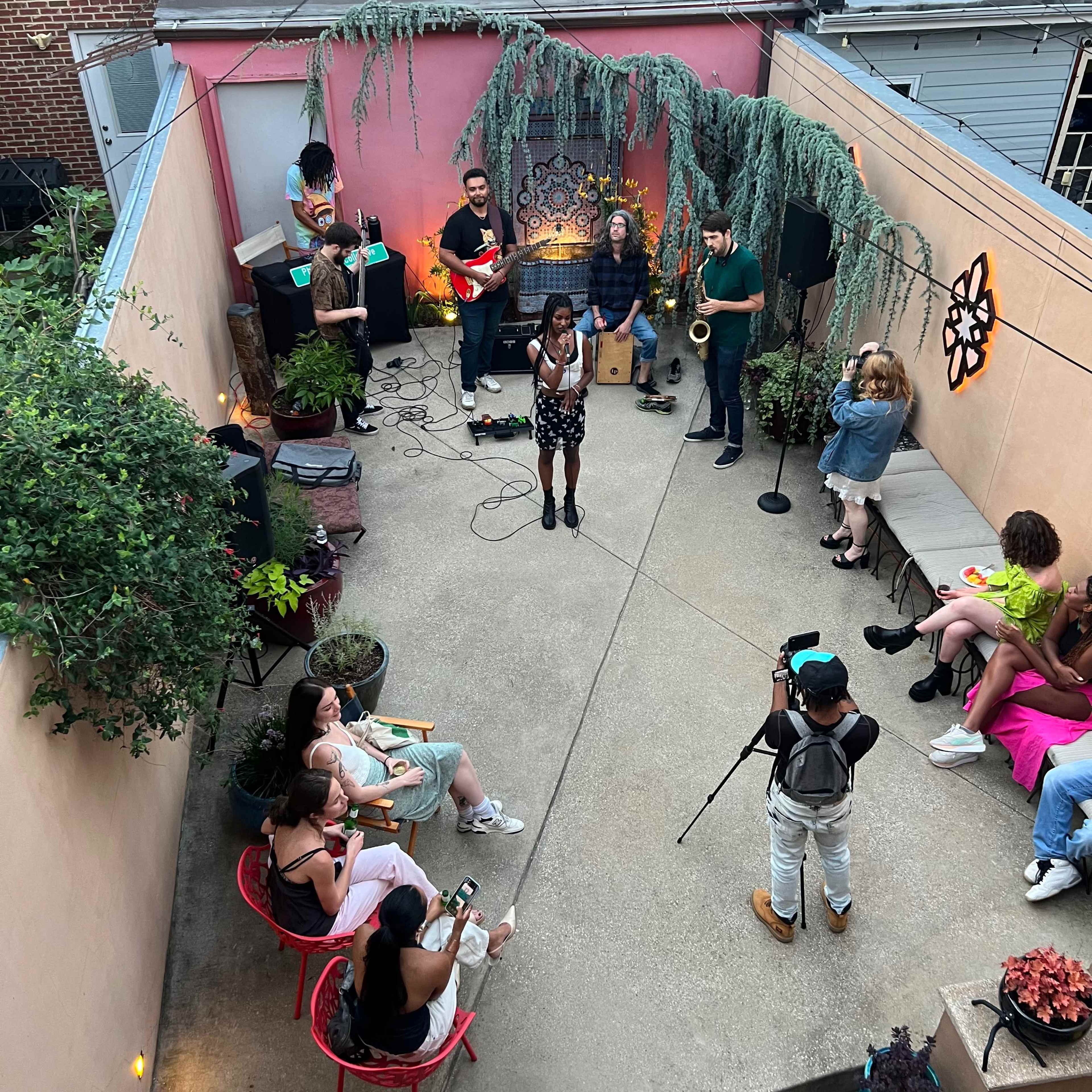 A group of musicians performs on a small outdoor stage while an audience sits on chairs and benches in a courtyard adorned with plants.