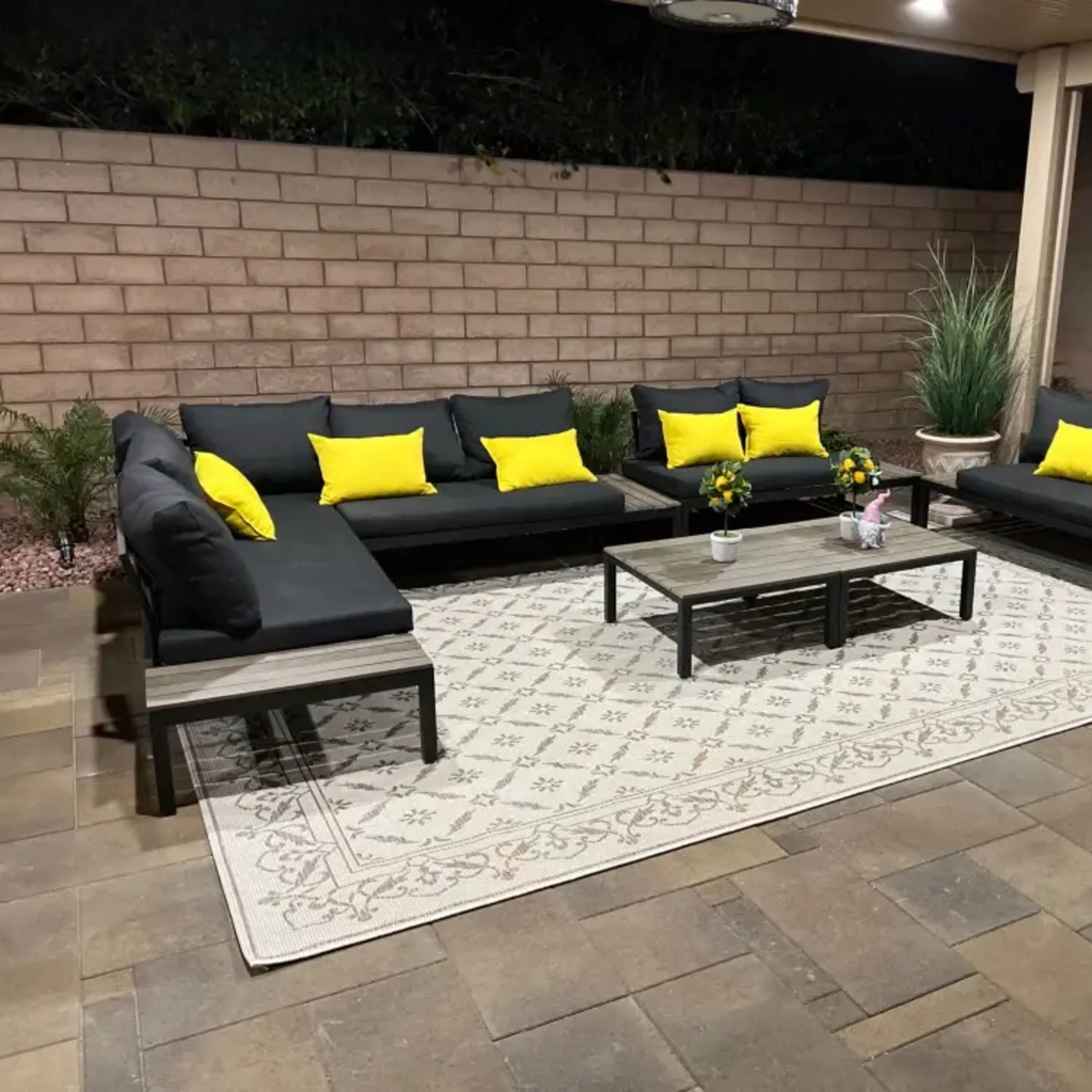 A patio area features a black sectional sofa with yellow cushions, a central table, and a large area rug on stone flooring.